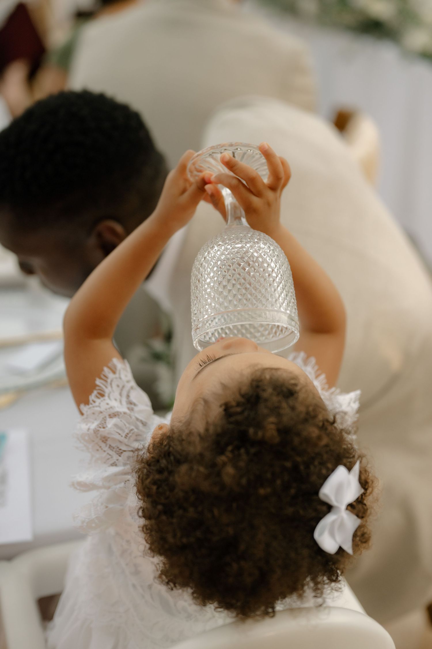 Two children in white outfits at a wedding reception, with a curly-haired girl holding up a glass vase.