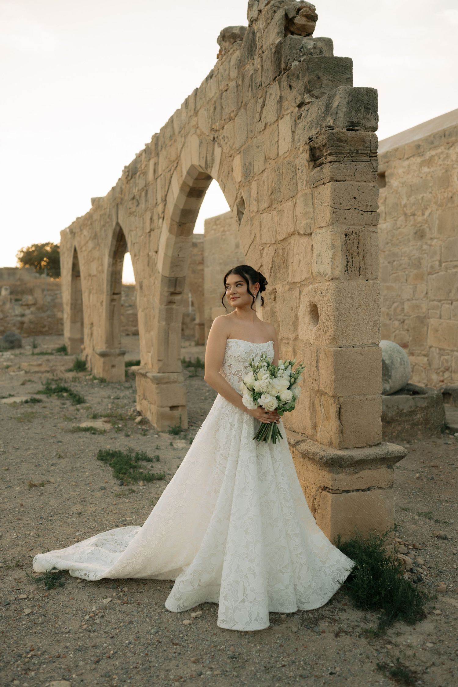 Bride in white strapless lace gown holds bouquet before ancient stone arches at sunset.