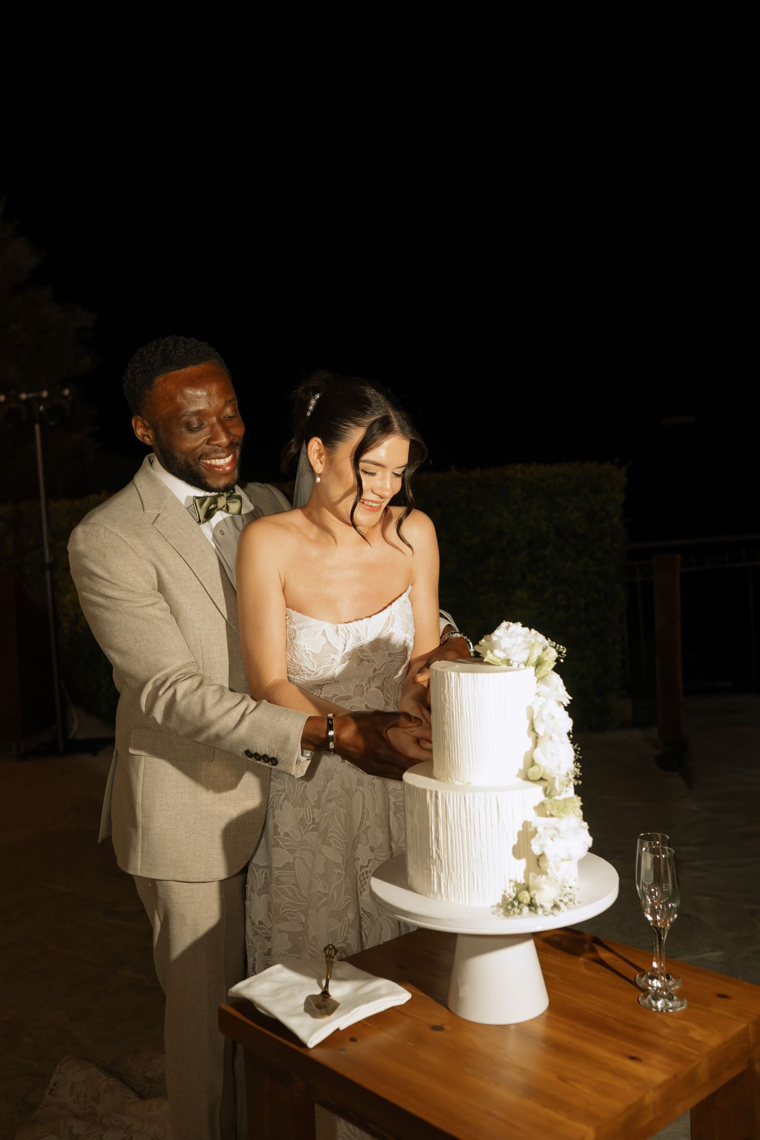 Couple cutting a white two-tier wedding cake outdoors at night, groom in tan suit, bride in strapless gown.