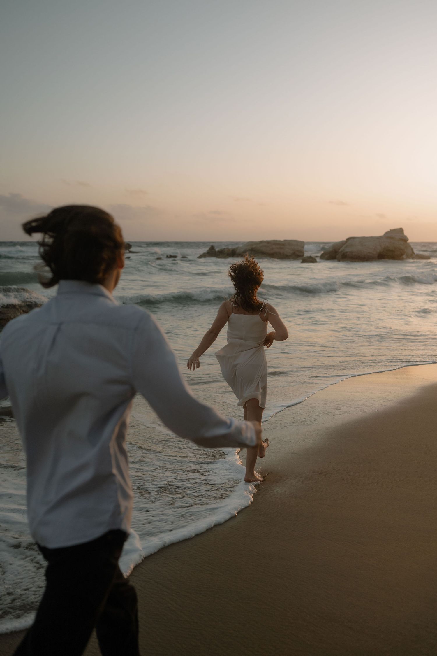 Couple running along shoreline at sunset, woman in white dress leads man in white shirt toward ocean waves.