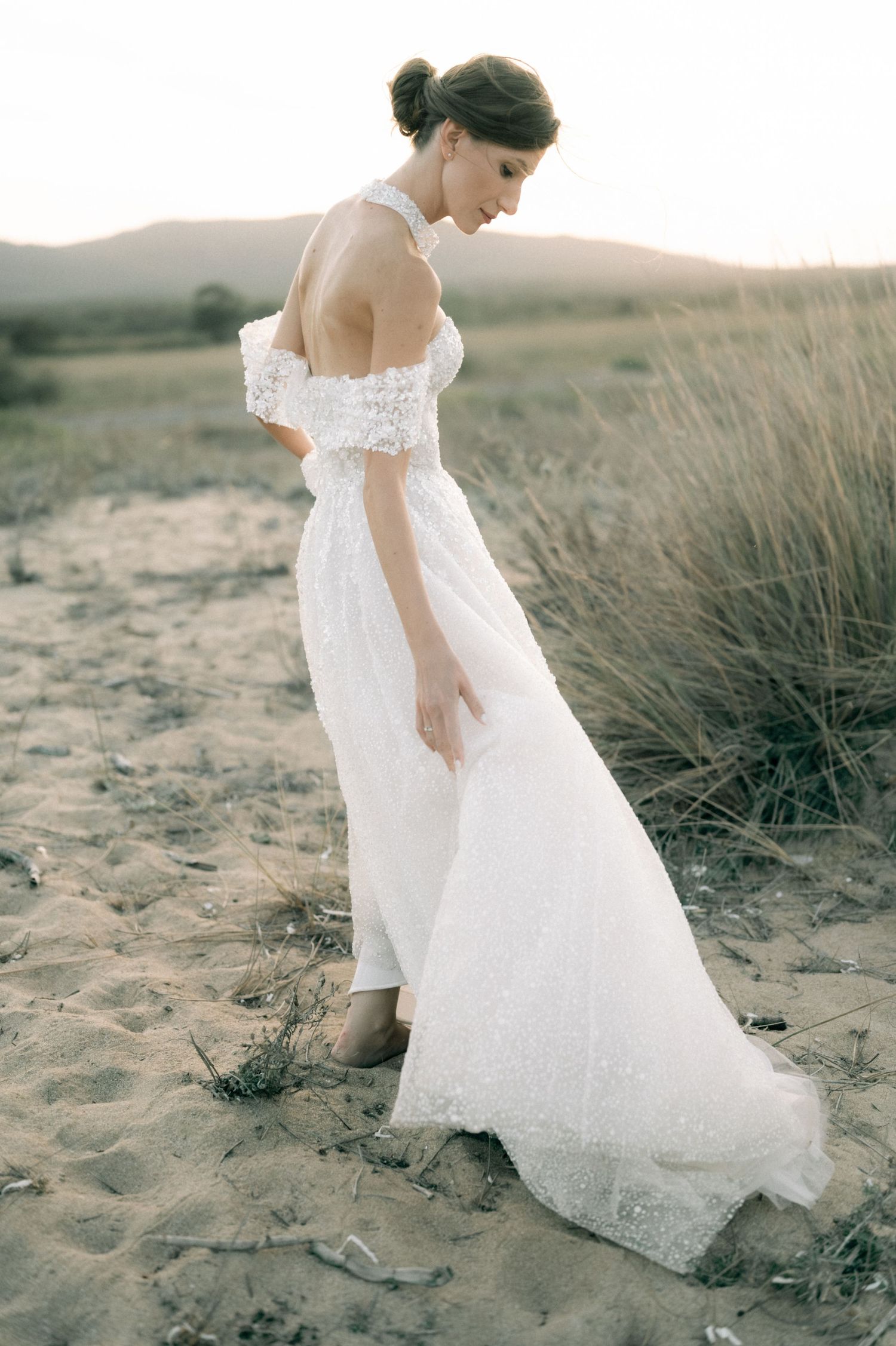 Bride in white off-shoulder lace wedding gown standing on sandy beach dunes at golden hour sunset.