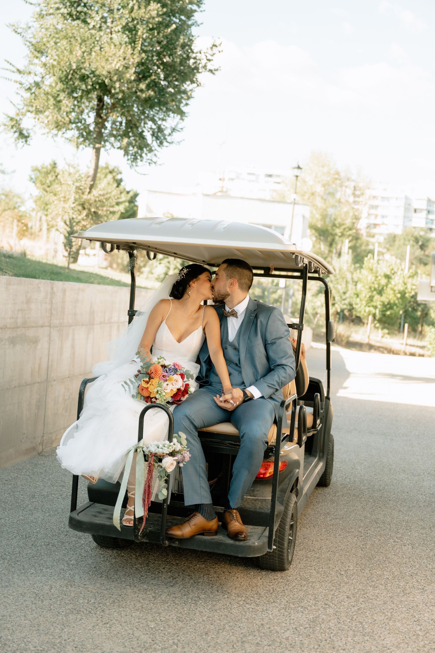A couple sits together in a golf cart at a wedding venue, with the bride's dress and bouquet visible.