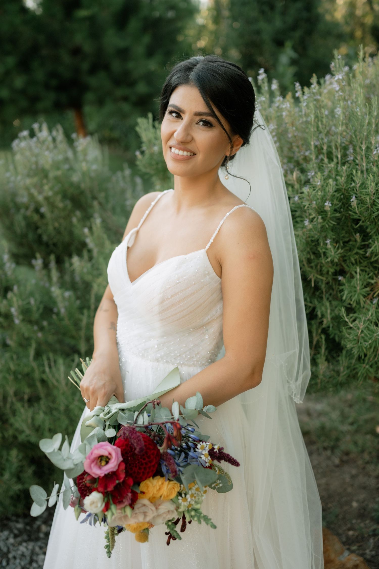 A bride in a white wedding dress holds a vibrant bouquet featuring burgundy and colorful flowers in an outdoor garden setting.