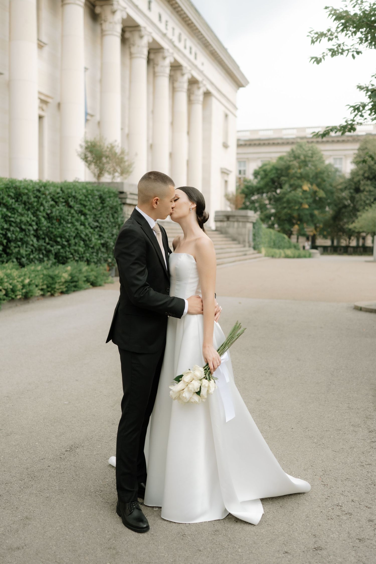 A romantic wedding portrait captured in front of a stately building with white columns and manicured hedges.