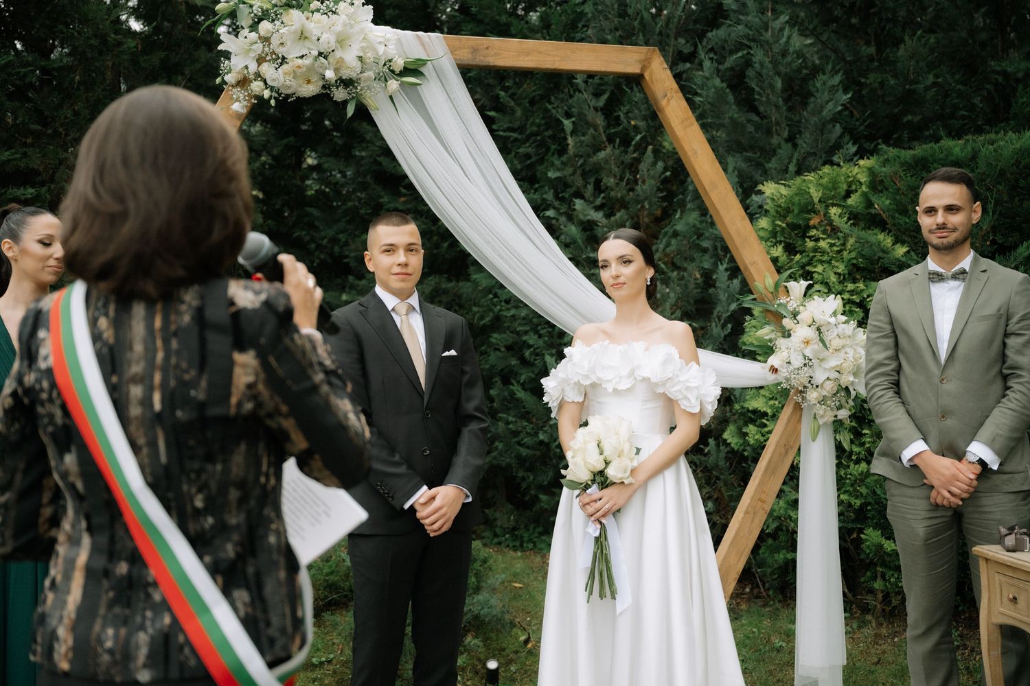 A wedding ceremony takes place under a wooden arch decorated with white fabric and floral arrangements in an outdoor garden setting.