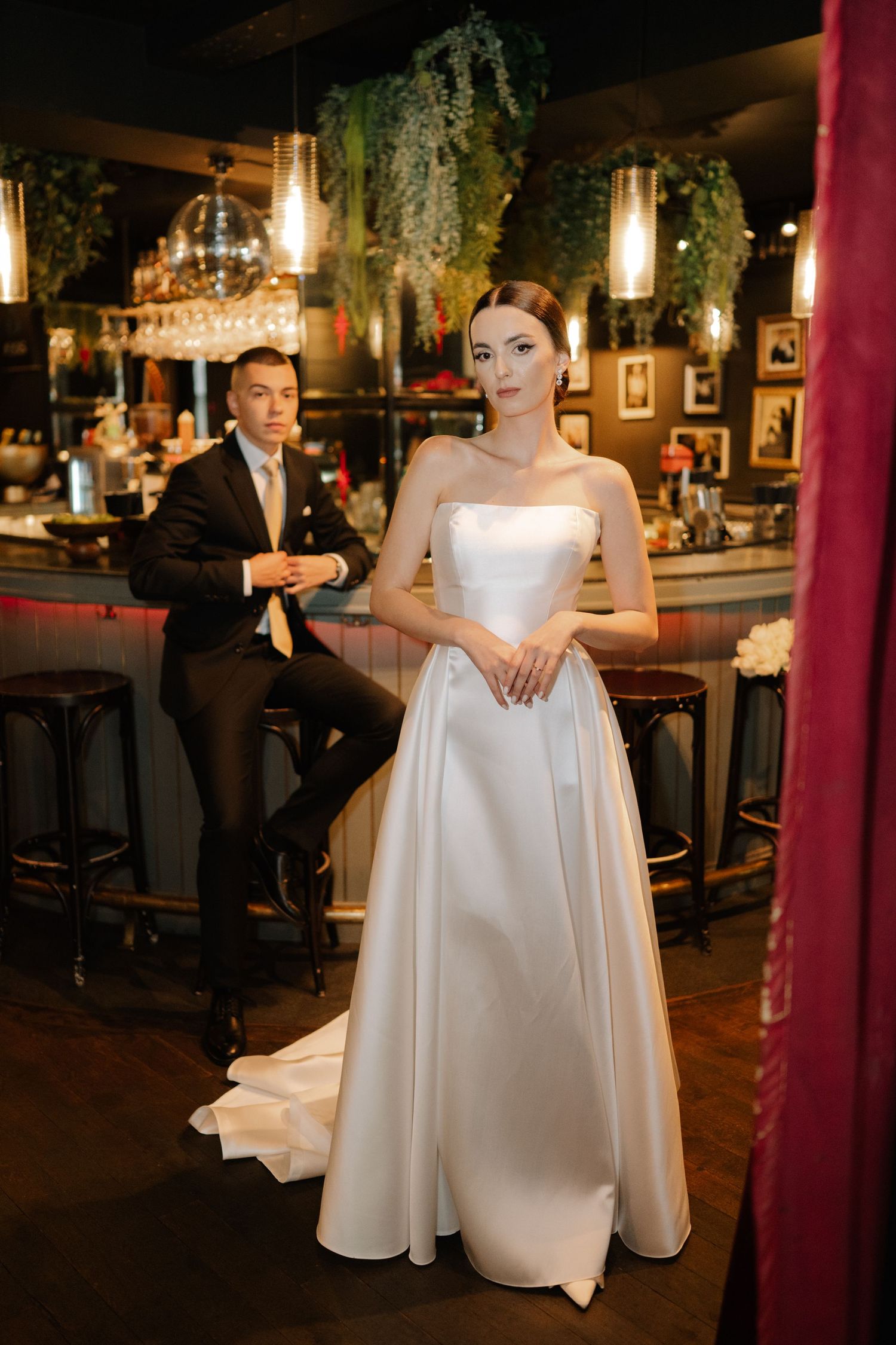Elegant wedding scene in a dimly lit bar with a bride in a white satin gown and someone seated at the counter.