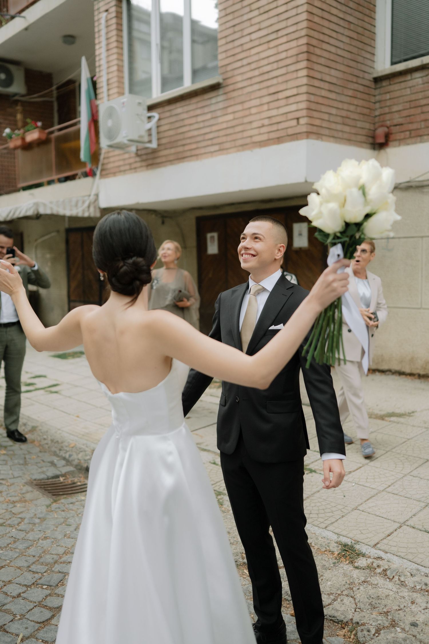 A bride in a white wedding dress holds a bouquet while walking with guests on a brick-paved courtyard.