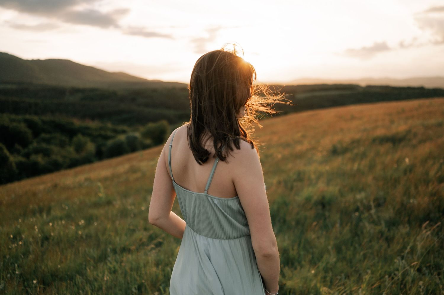 Woman in gray sundress standing in golden meadow at sunset with rolling hills and dramatic sky backdrop.