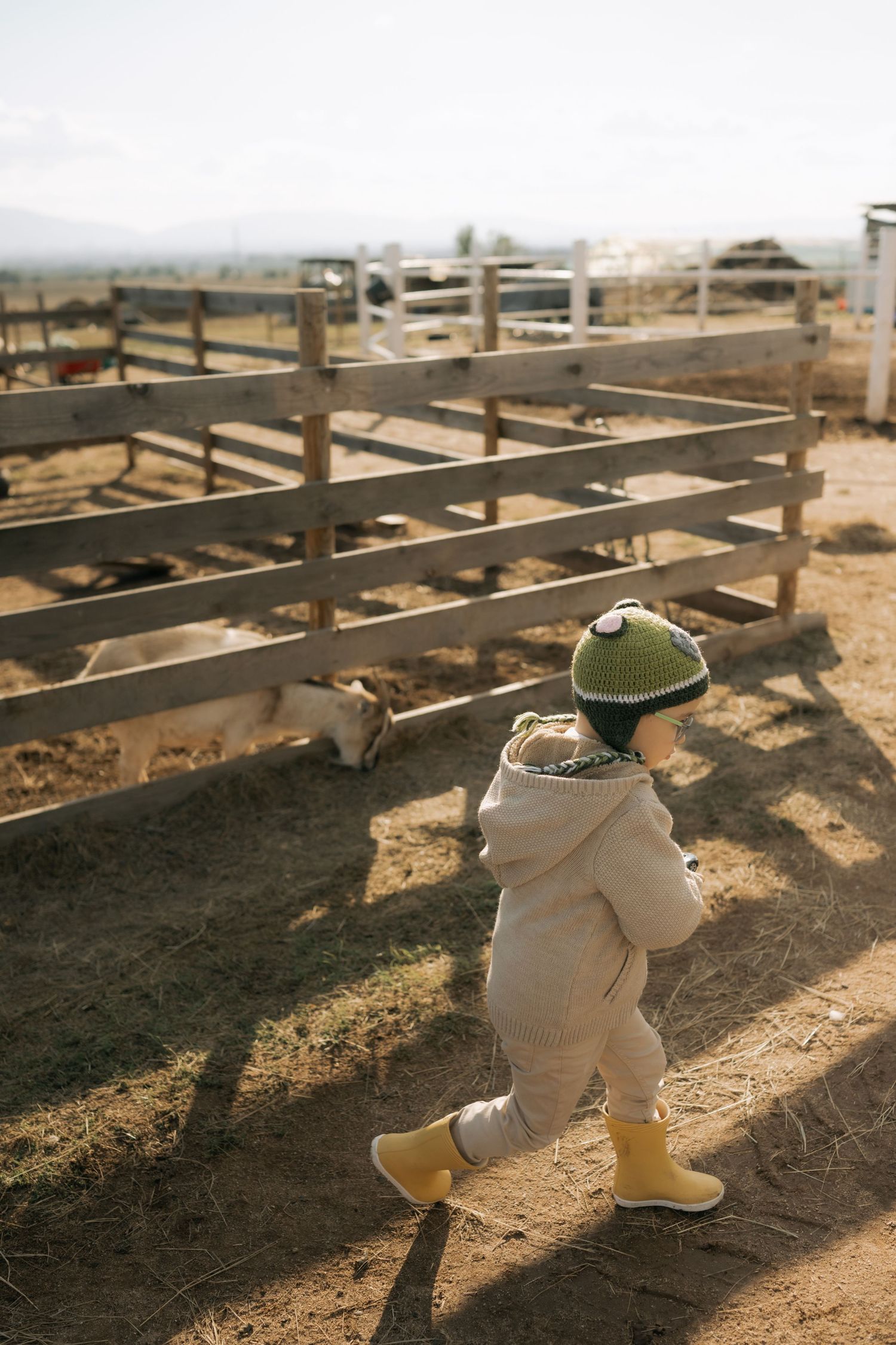 A toddler in a white jacket and hat crawls under wooden bleachers casting long shadows on dirt ground.