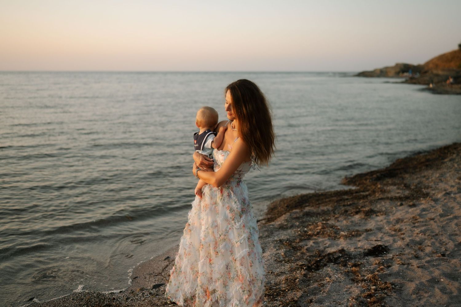 A figure in a flowing dress stands on a rocky beach at sunset against a serene coastal landscape.