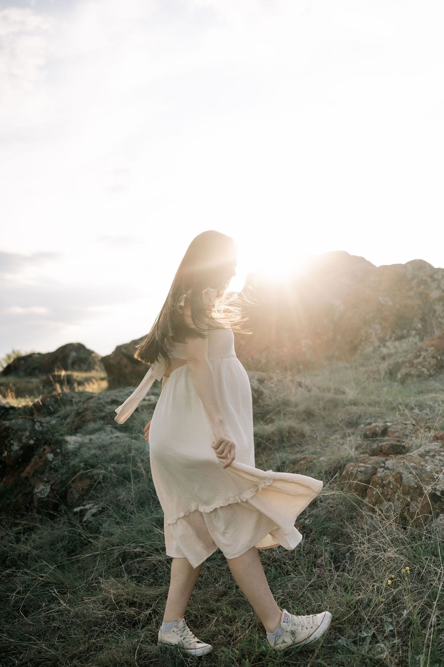 A figure in a flowing white dress walks through a sunlit mountainous landscape at golden hour.