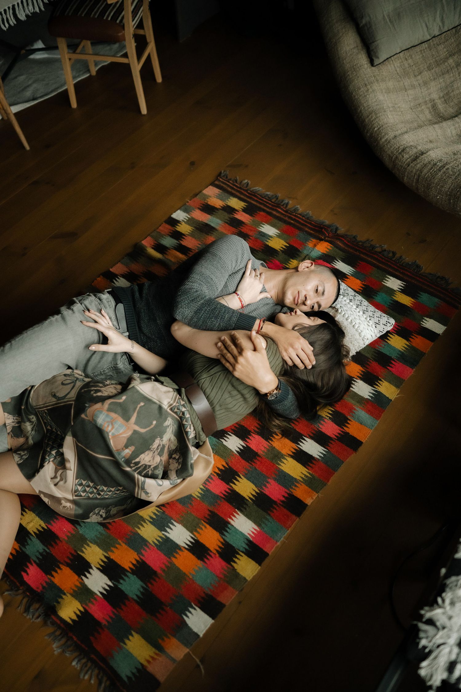 A couple lies together on a colorful patchwork rug in a dimly lit room creating an intimate moment.
