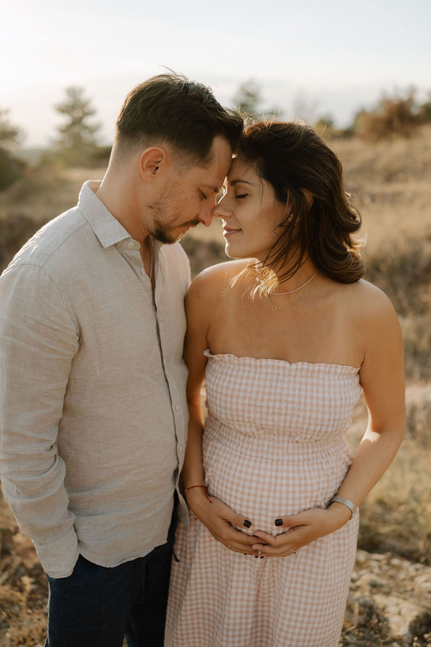 A tender maternity photo showing a couple in a field at sunset, with the woman wearing a gingham dress and cradling her belly.