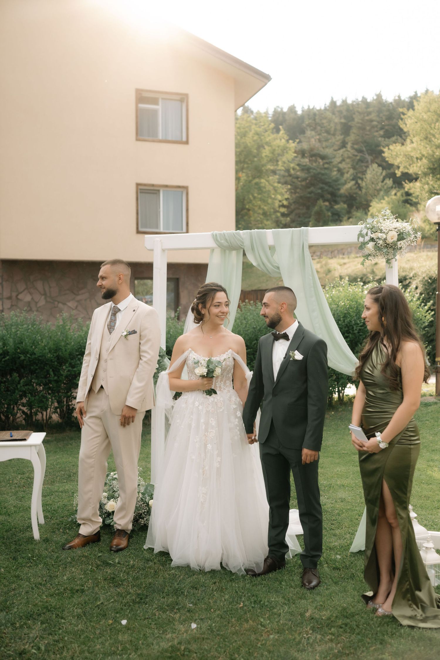 Wedding party gathered on a lawn with decorative white backdrop and greenery for outdoor celebration photos.