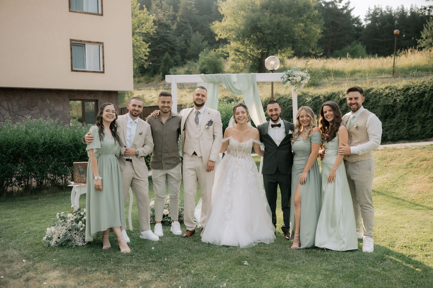 Wedding party in light sage green and beige attire poses together on a grassy hillside during a summer celebration.