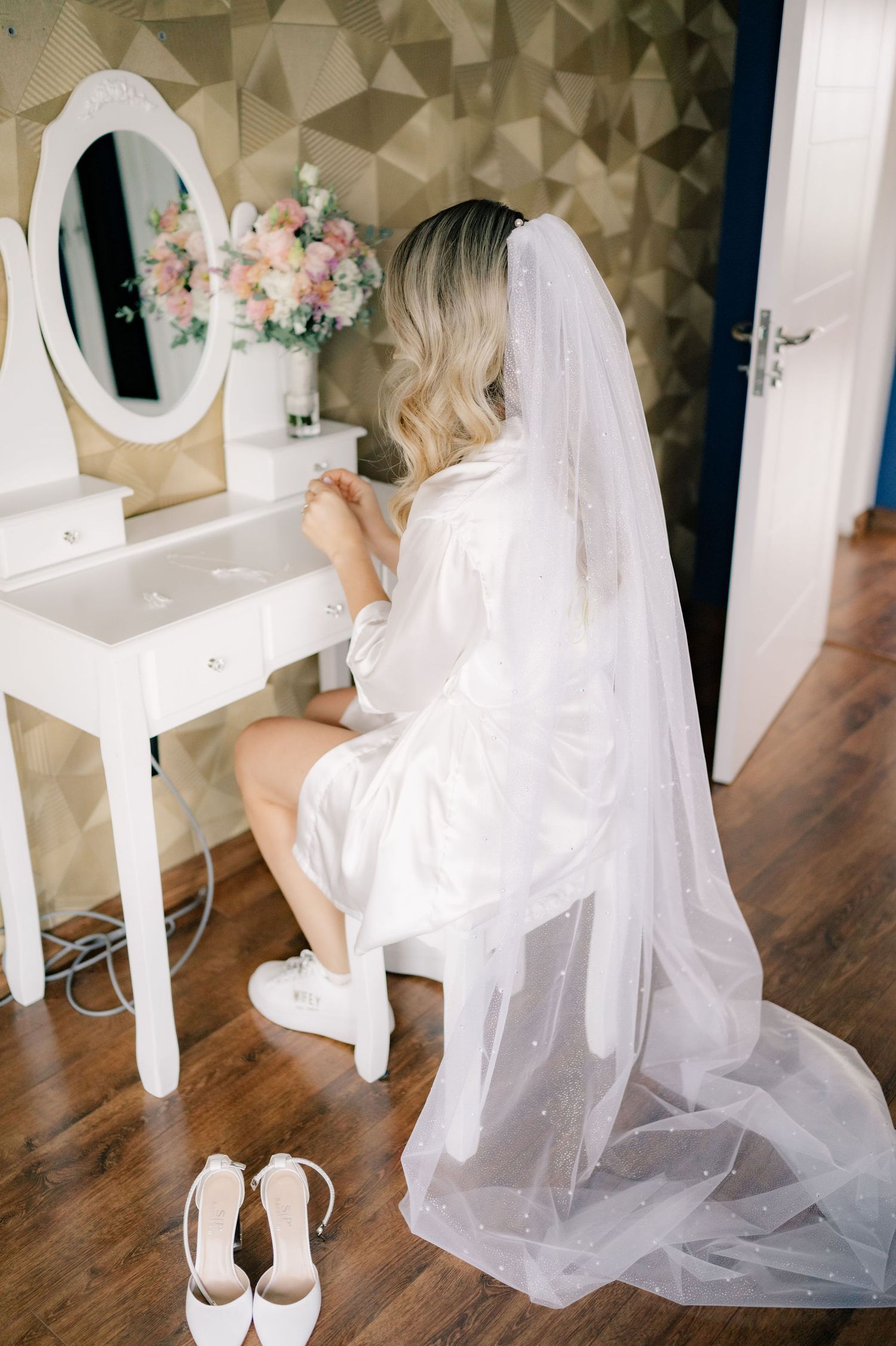A white vanity table with an oval mirror and floral arrangement sits against a textured wall in a bridal preparation room.