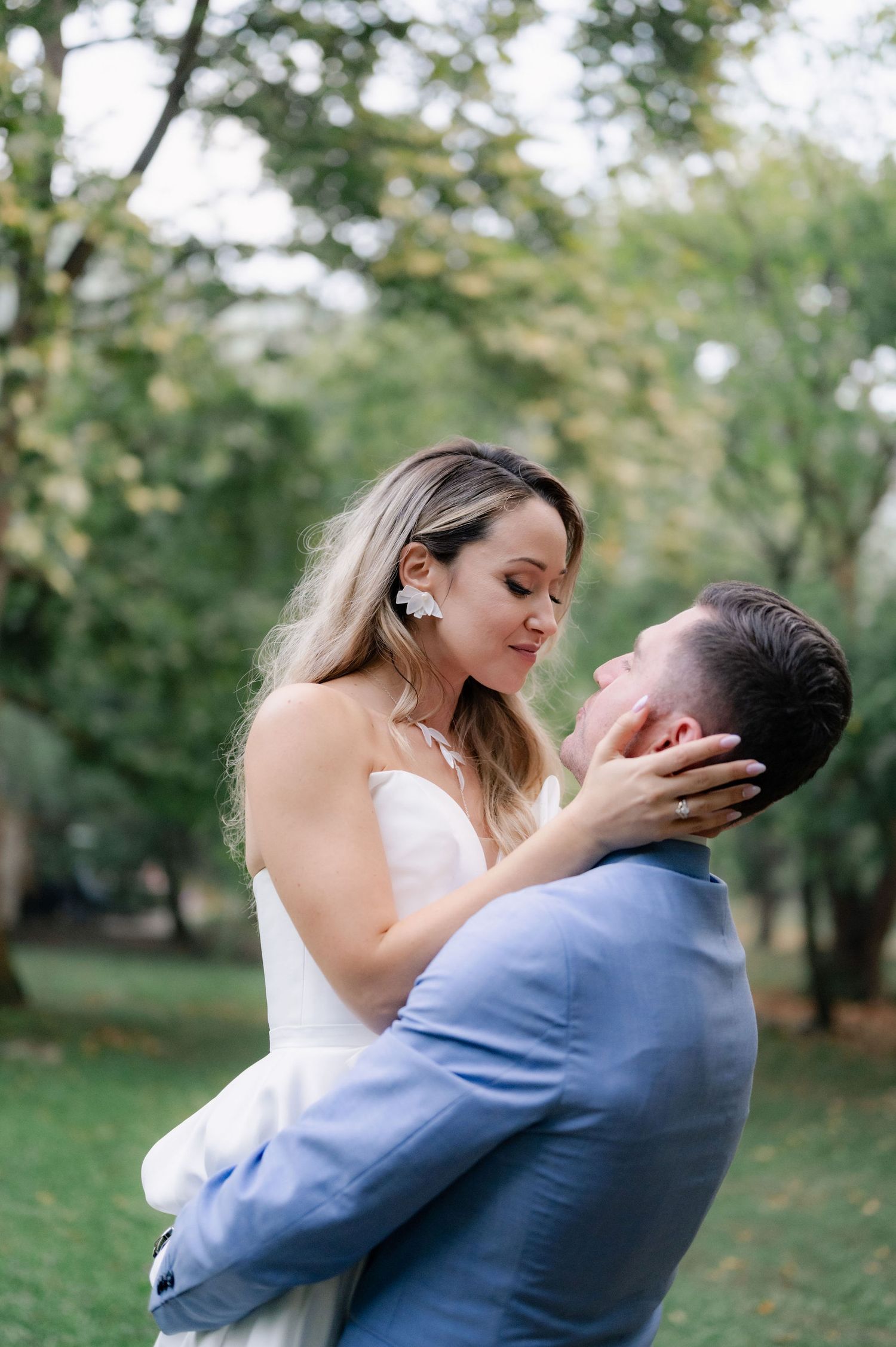 A romantic wedding couple embraces in a park with autumn trees in the background.