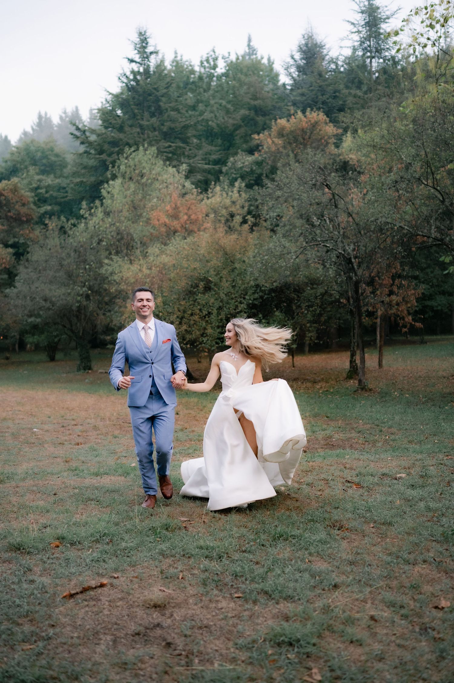 A couple in wedding attire walks through a wooded area with evergreen trees and autumn foliage in the background.