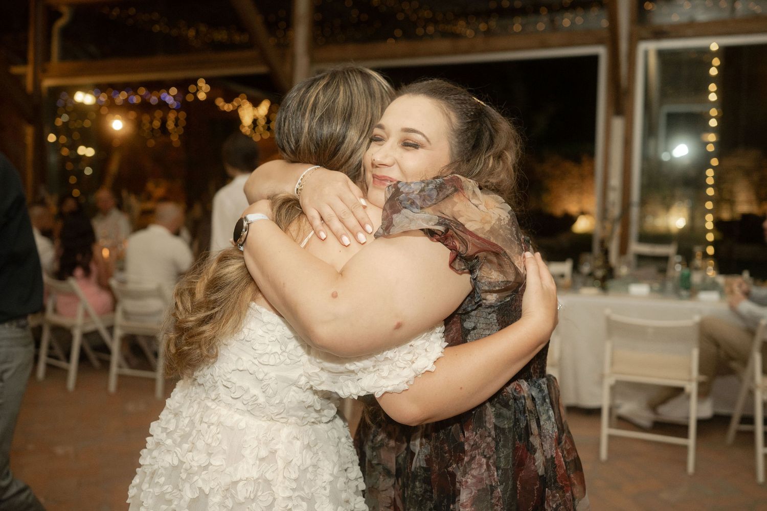 Two people share a warm embrace at an evening wedding reception with string lights and white tables in the background.