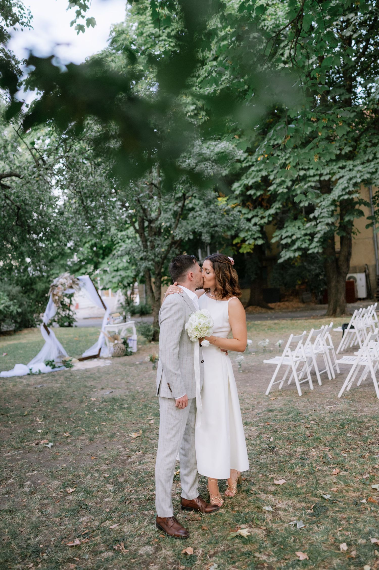 A romantic outdoor wedding ceremony setup with white chairs on a grassy lawn surrounded by lush green trees.