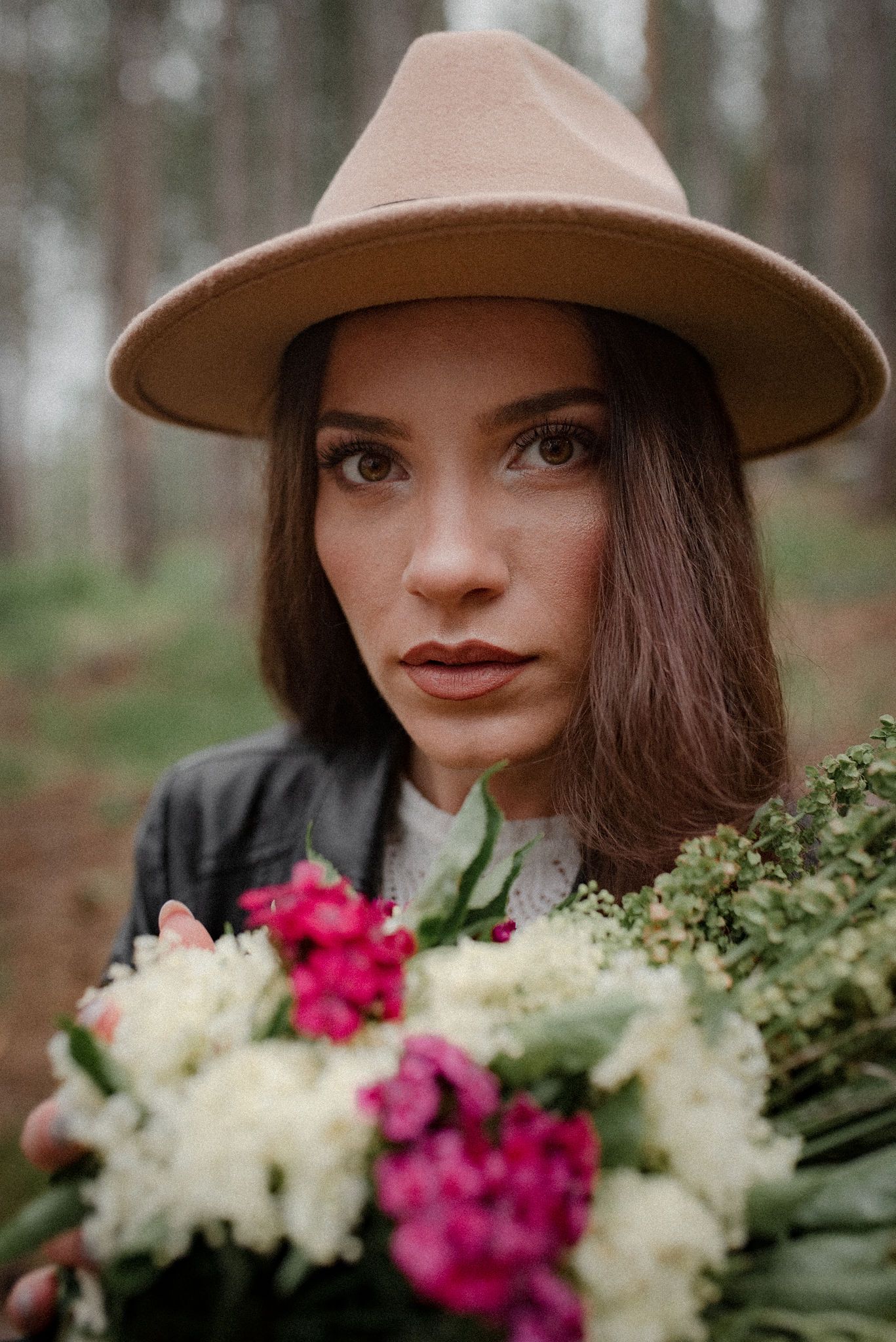 A person in a brown felt hat holds a bouquet of pink and white flowers while standing in a forest setting.