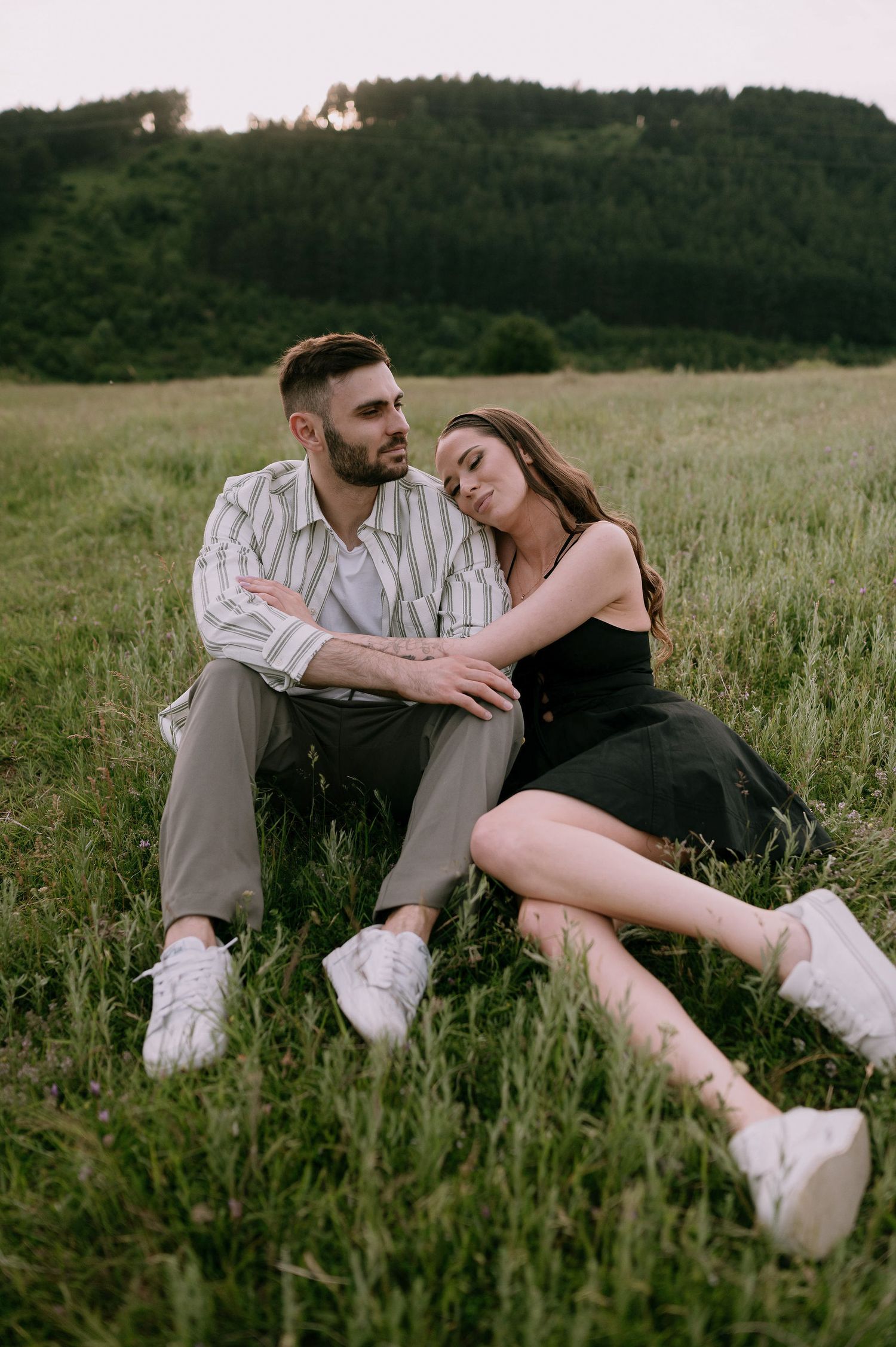 A couple relaxes together in a grassy meadow at sunset wearing casual clothes and white sneakers.