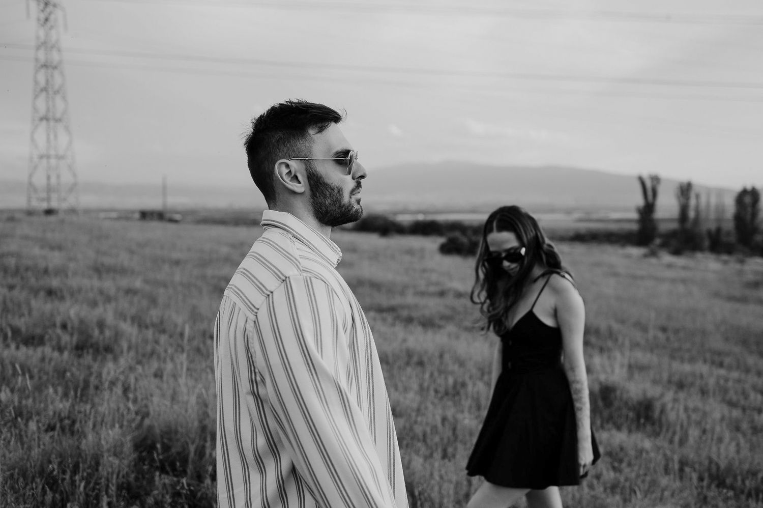 Black and white photo of two people standing in a field with power lines and mountains visible in the distance.