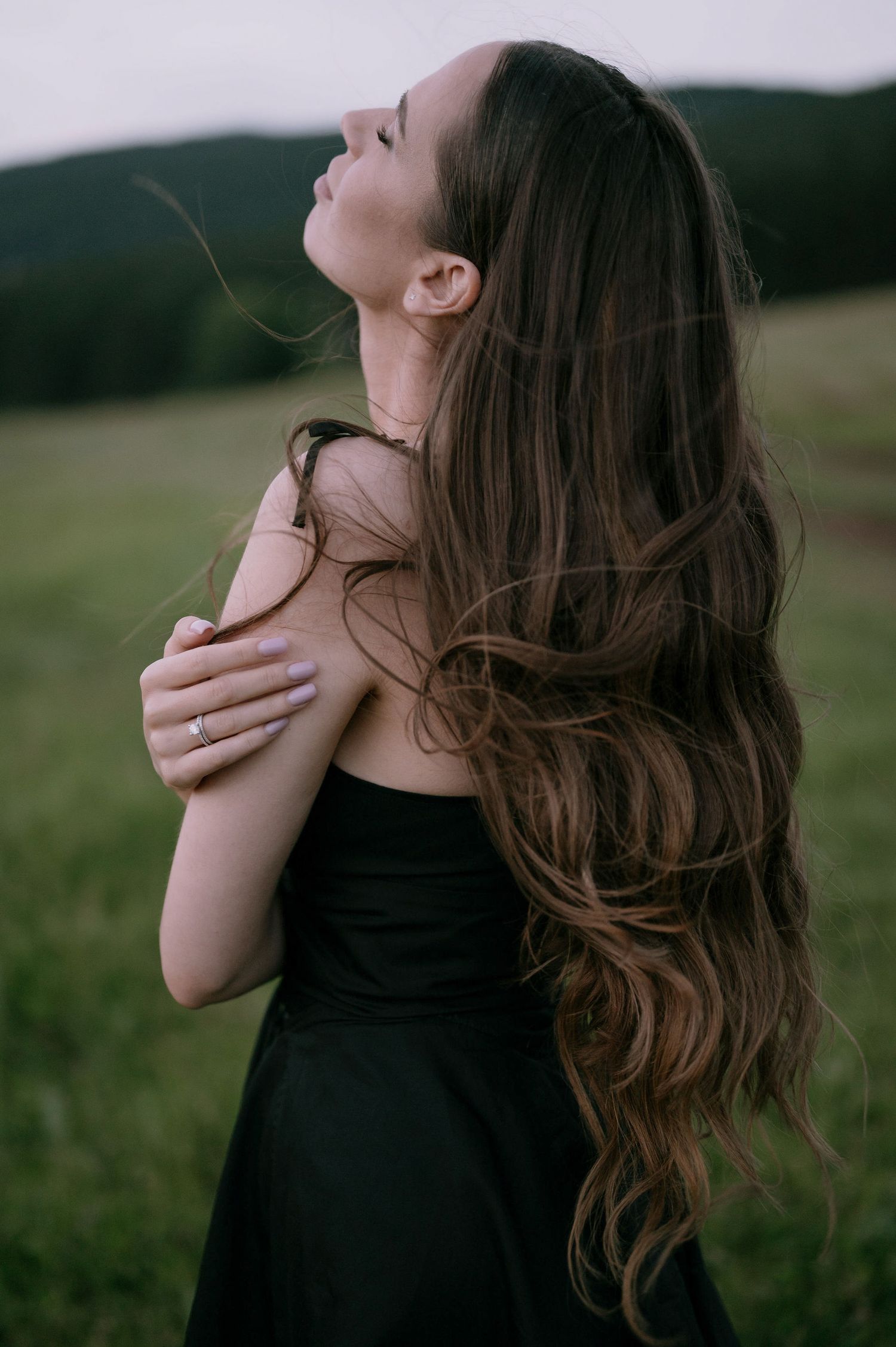 A person with long wavy brown hair wearing a black dress stands in a meadow with eyes closed and hand on shoulder.
