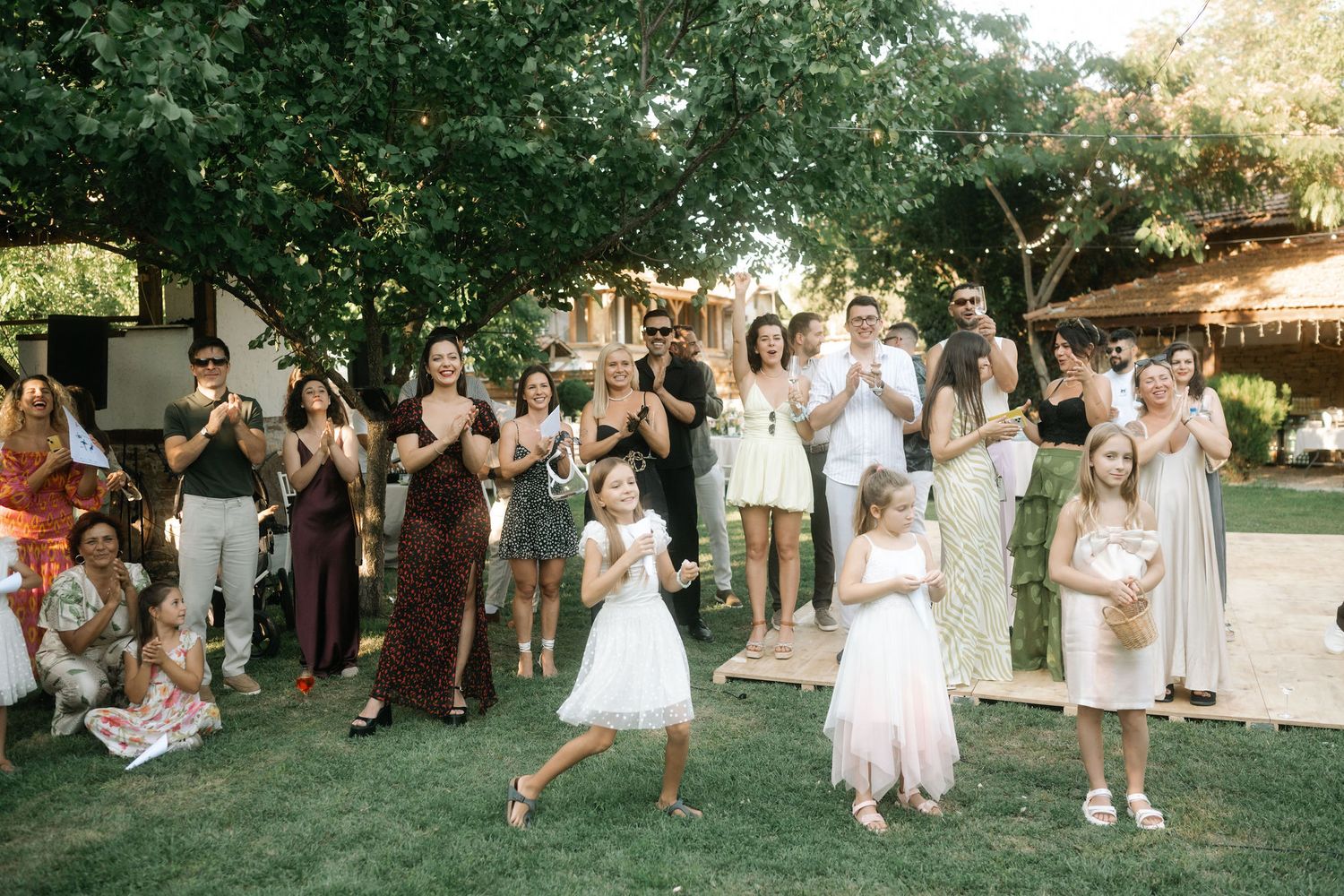 Wedding guests dance and celebrate outdoors at a summer garden reception under string lights and trees.