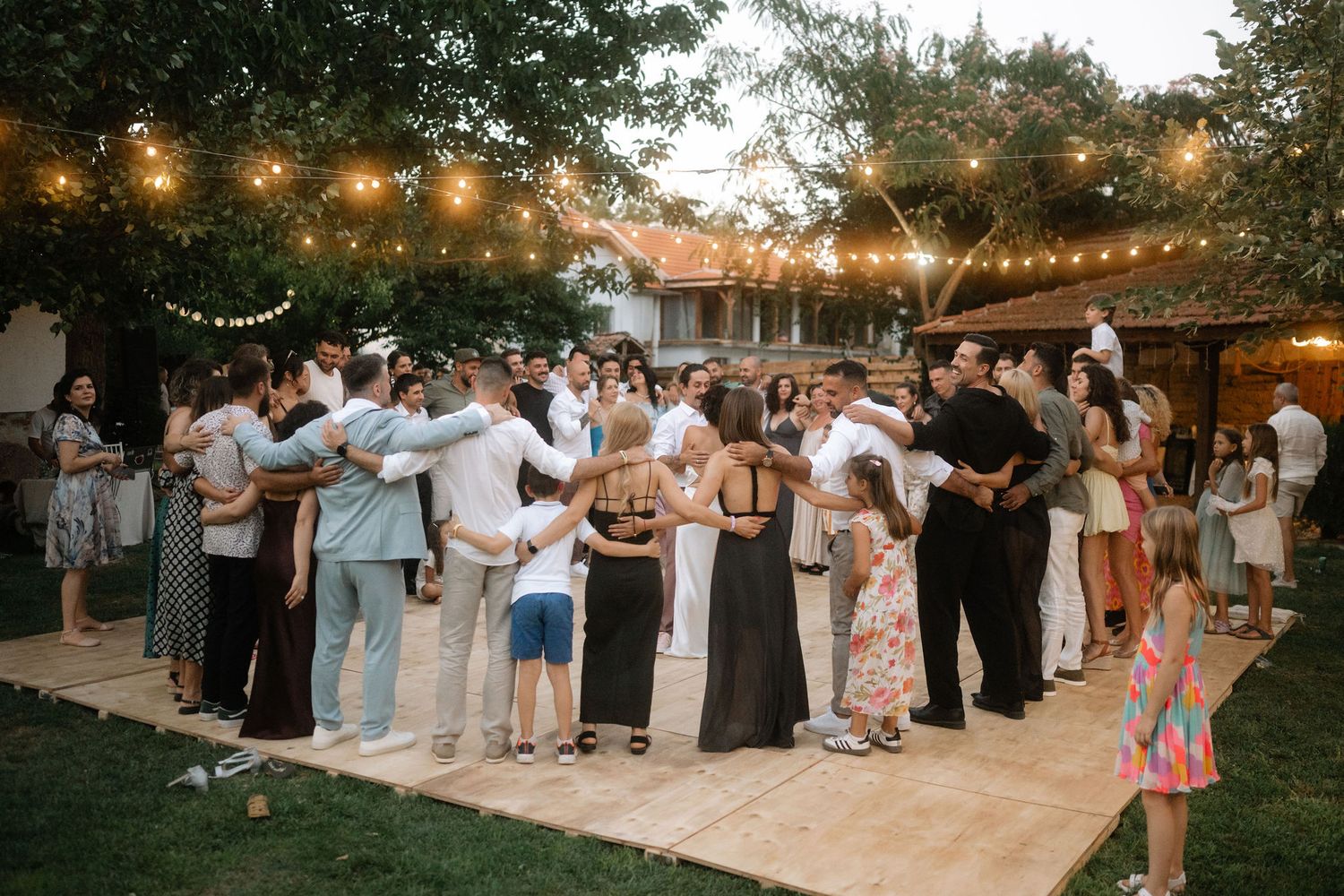Wedding guests dance together on an outdoor patio decorated with string lights during sunset reception celebration.