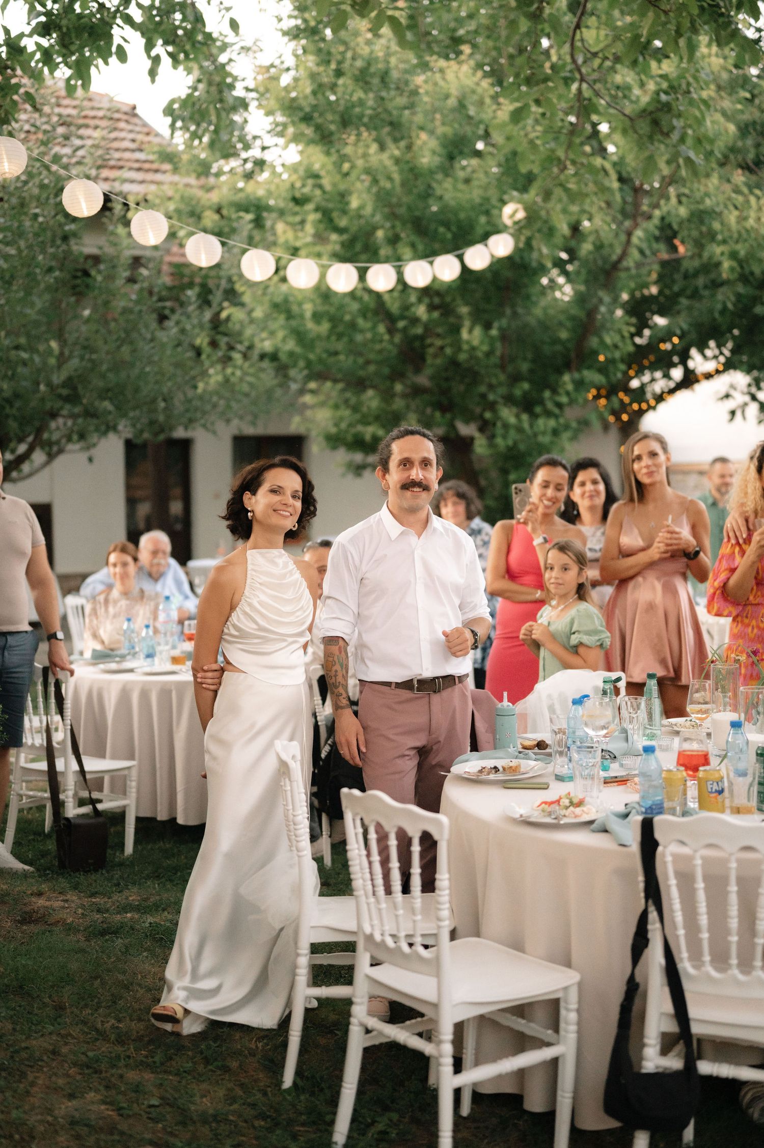 Guests enjoying an elegant outdoor reception dinner under string lights and trees with white table settings and decor.