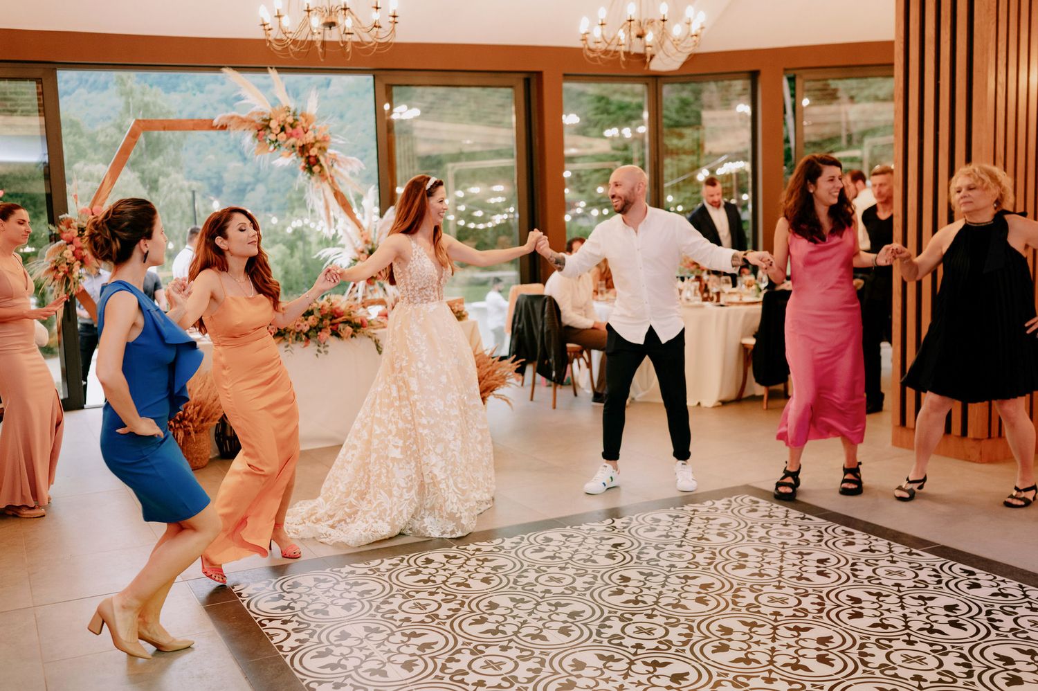 Wedding guests dance on a patterned floor under elegant chandeliers at an indoor reception venue.