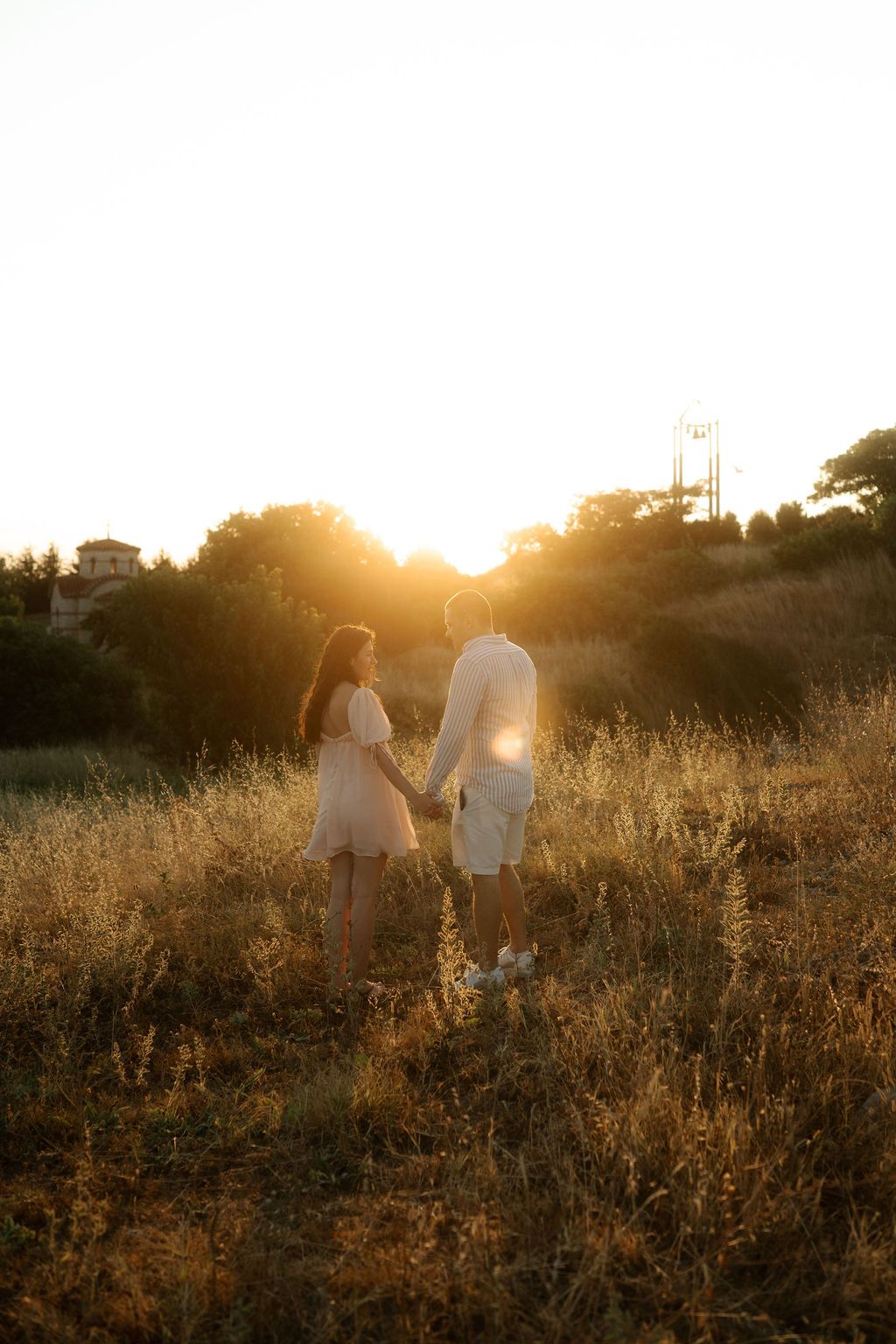 A couple holds hands while walking through a golden field at sunset, backlit by warm evening light.