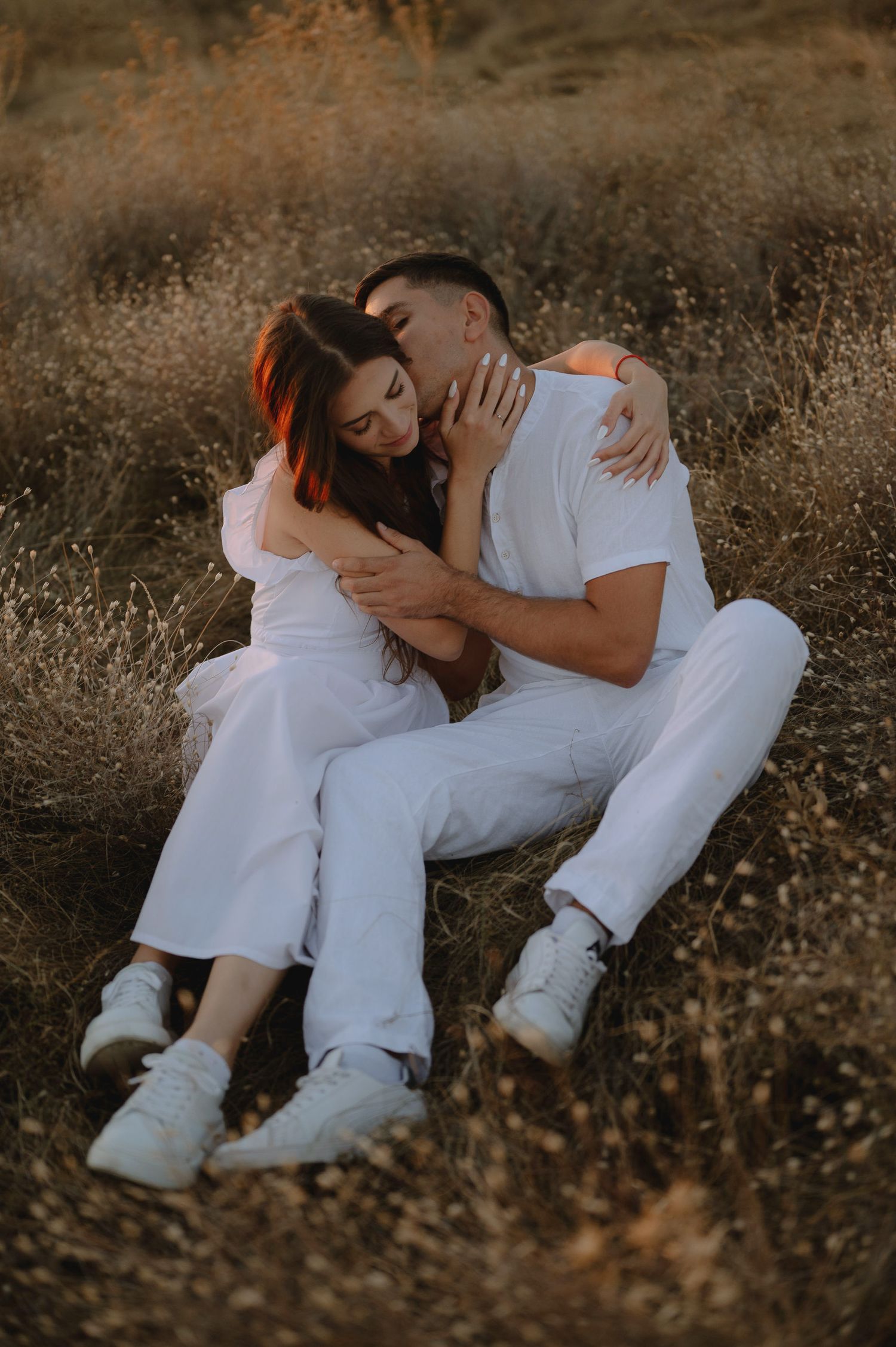A couple in white outfits embraces intimately while laying in a rustic outdoor setting during golden hour.