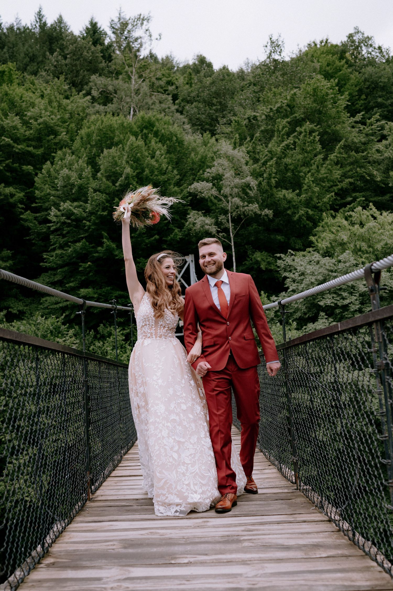 A couple in wedding attire walks together on a wooden suspension bridge surrounded by lush green forest.