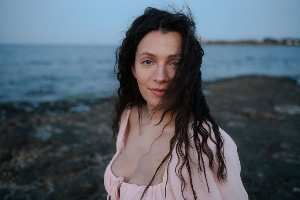 A person with dark hair poses by the ocean at dusk wearing a light colored top.