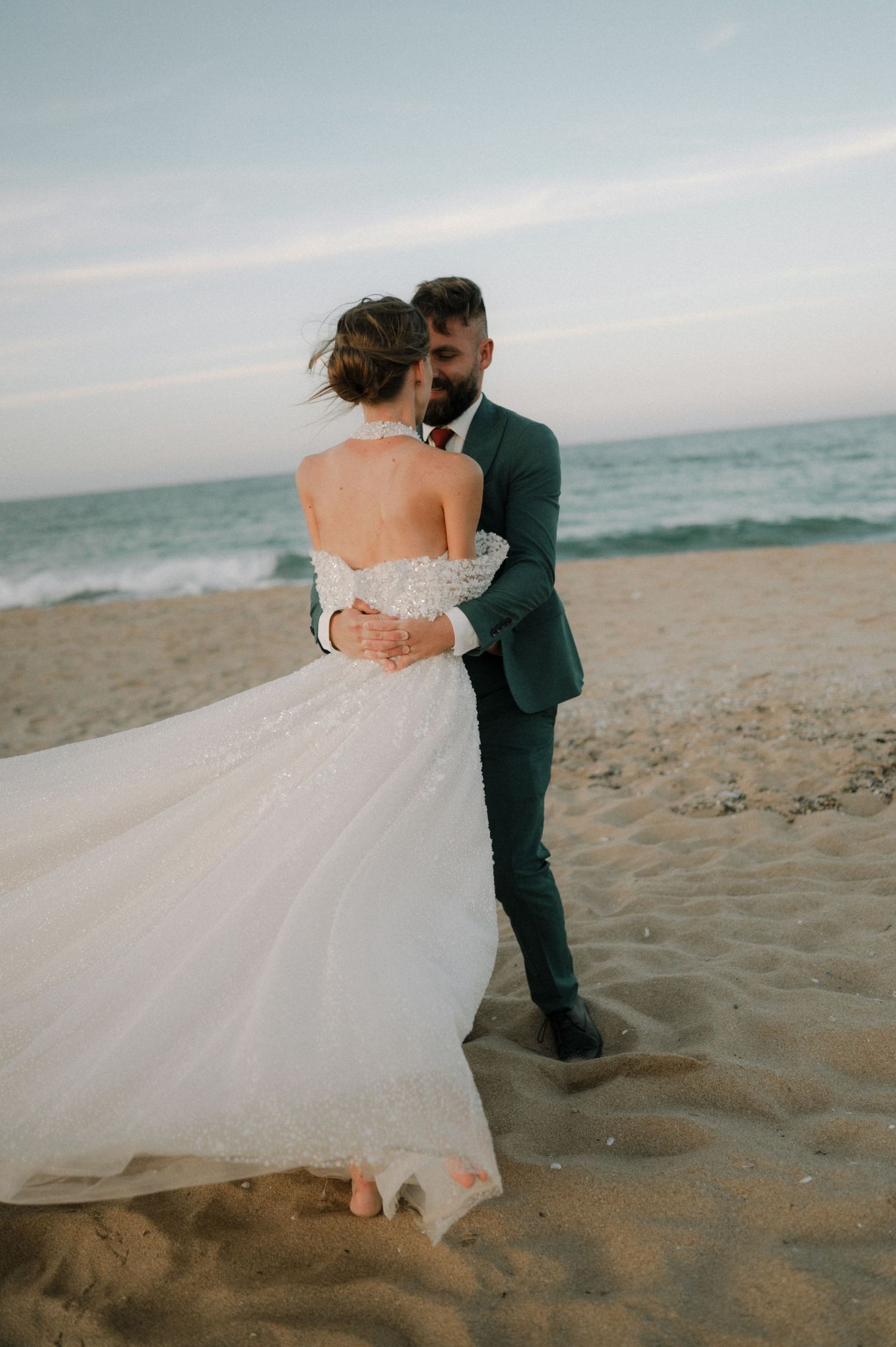 Bride and groom embrace on sandy beach at sunset, she wears a flowing white gown as waves crash behind them.