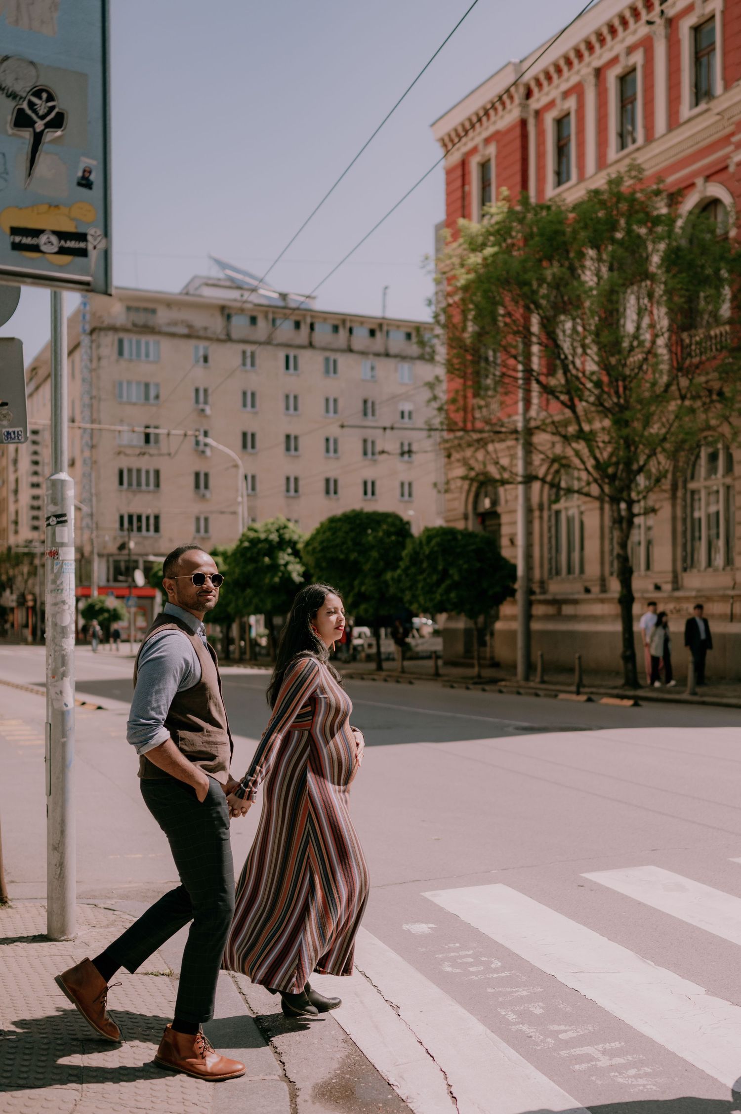 Pregnant couple holding hands crossing a sunny urban street, woman in striped maxi dress, classic buildings in background.