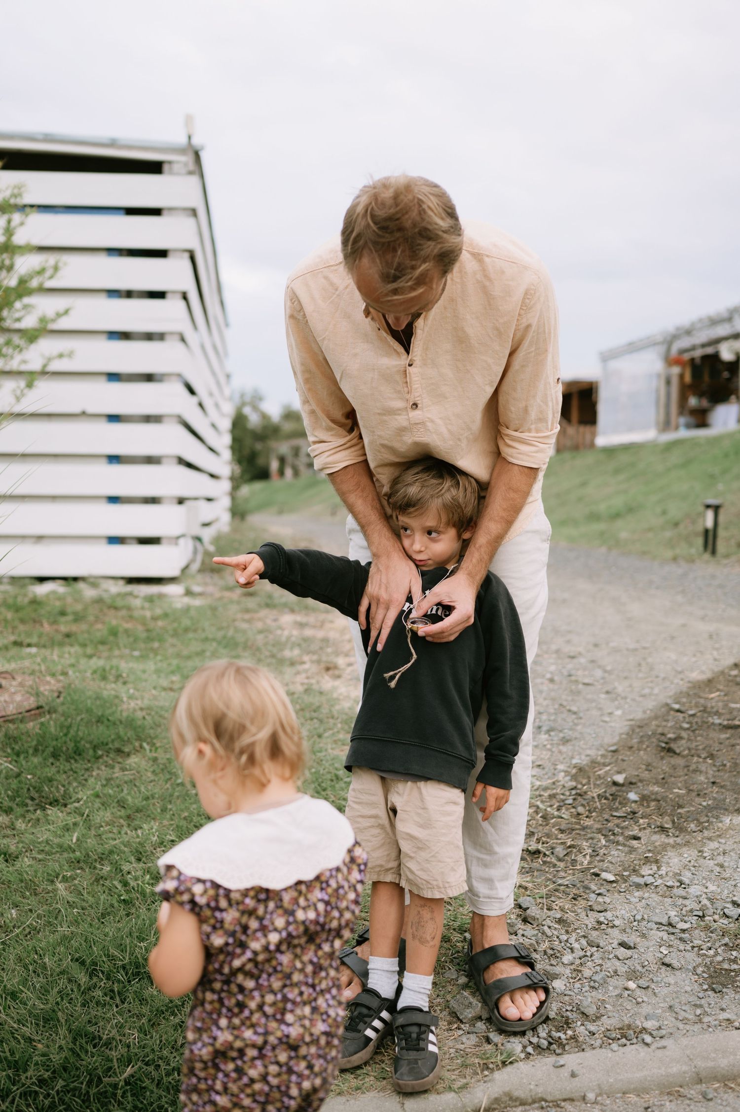 A man bends down toward two young children on a gravel path near farm buildings outdoors.