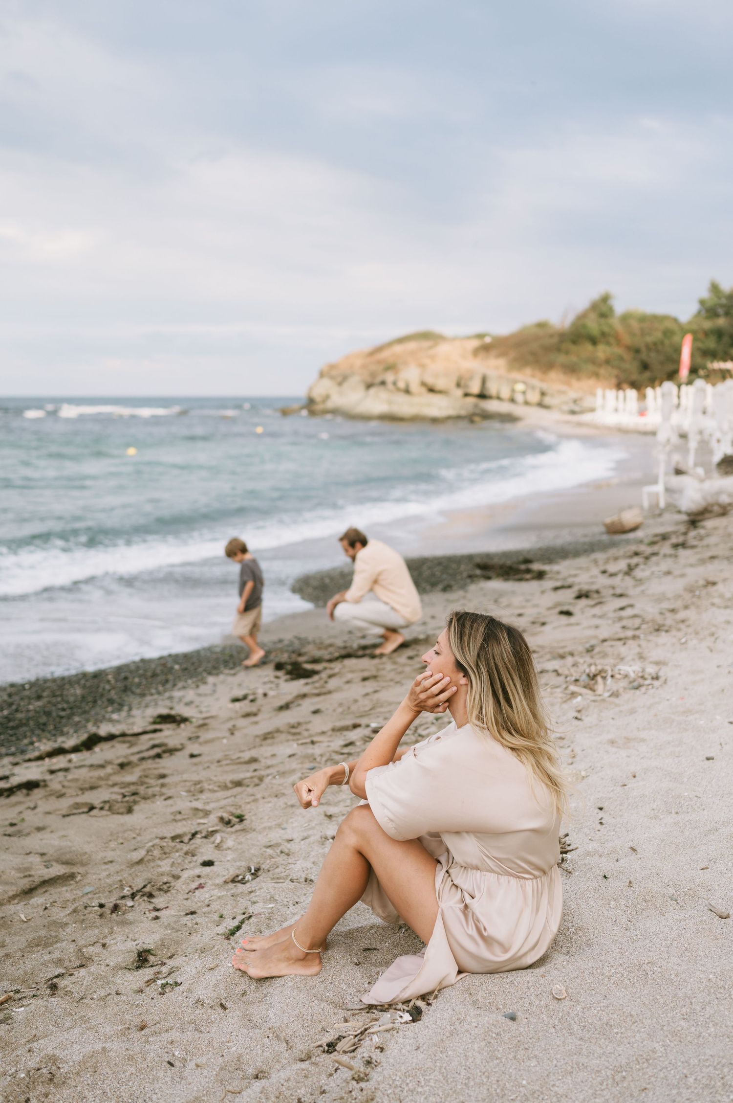 Woman in beige dress sitting on sandy beach with two people walking near ocean waves and rocky coastline.
