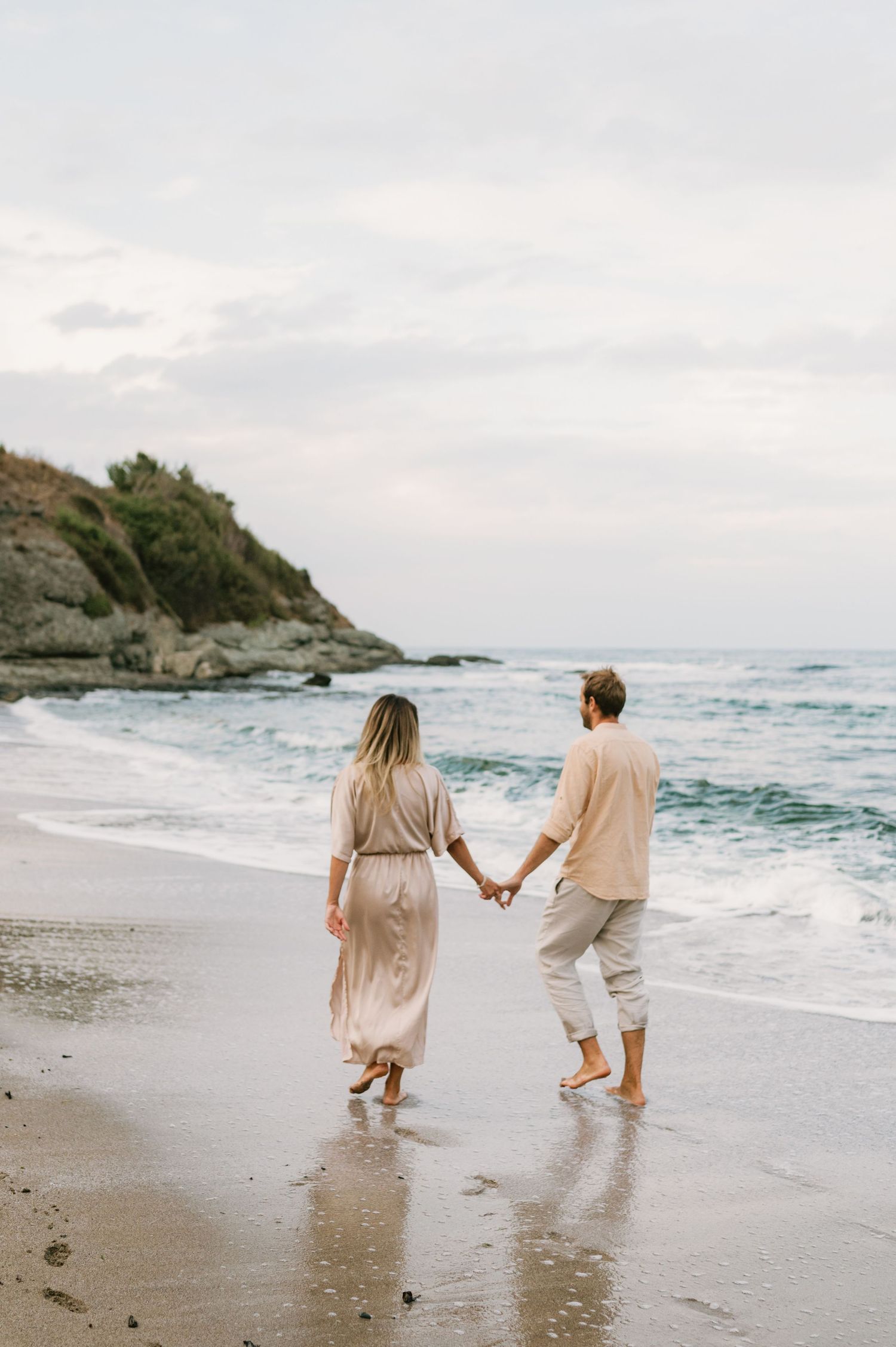 Couple holding hands walking barefoot on a sandy beach near ocean waves at dusk in neutral linen outfits.