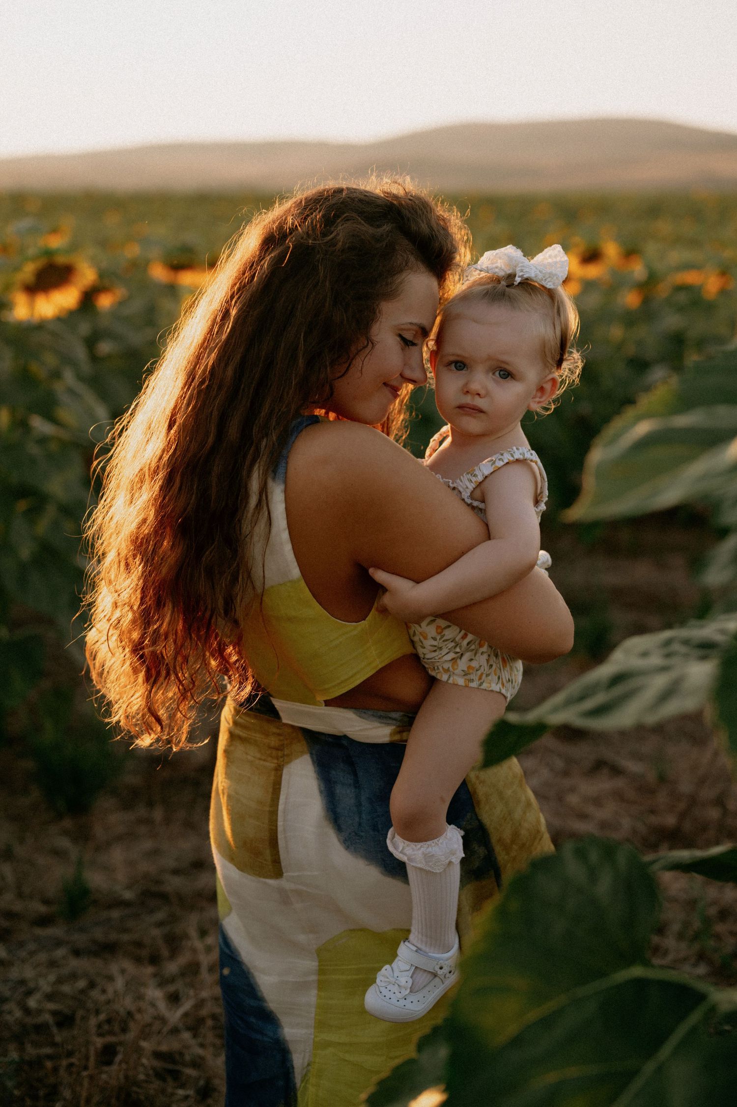 A mother holds her baby girl in a sunflower field at golden hour, both wearing summer outfits.