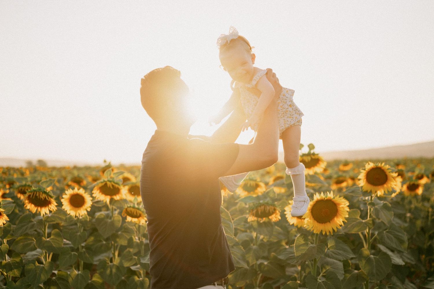 Father lifting baby girl in sunflower field at golden hour sunset, joyful family moment outdoors.