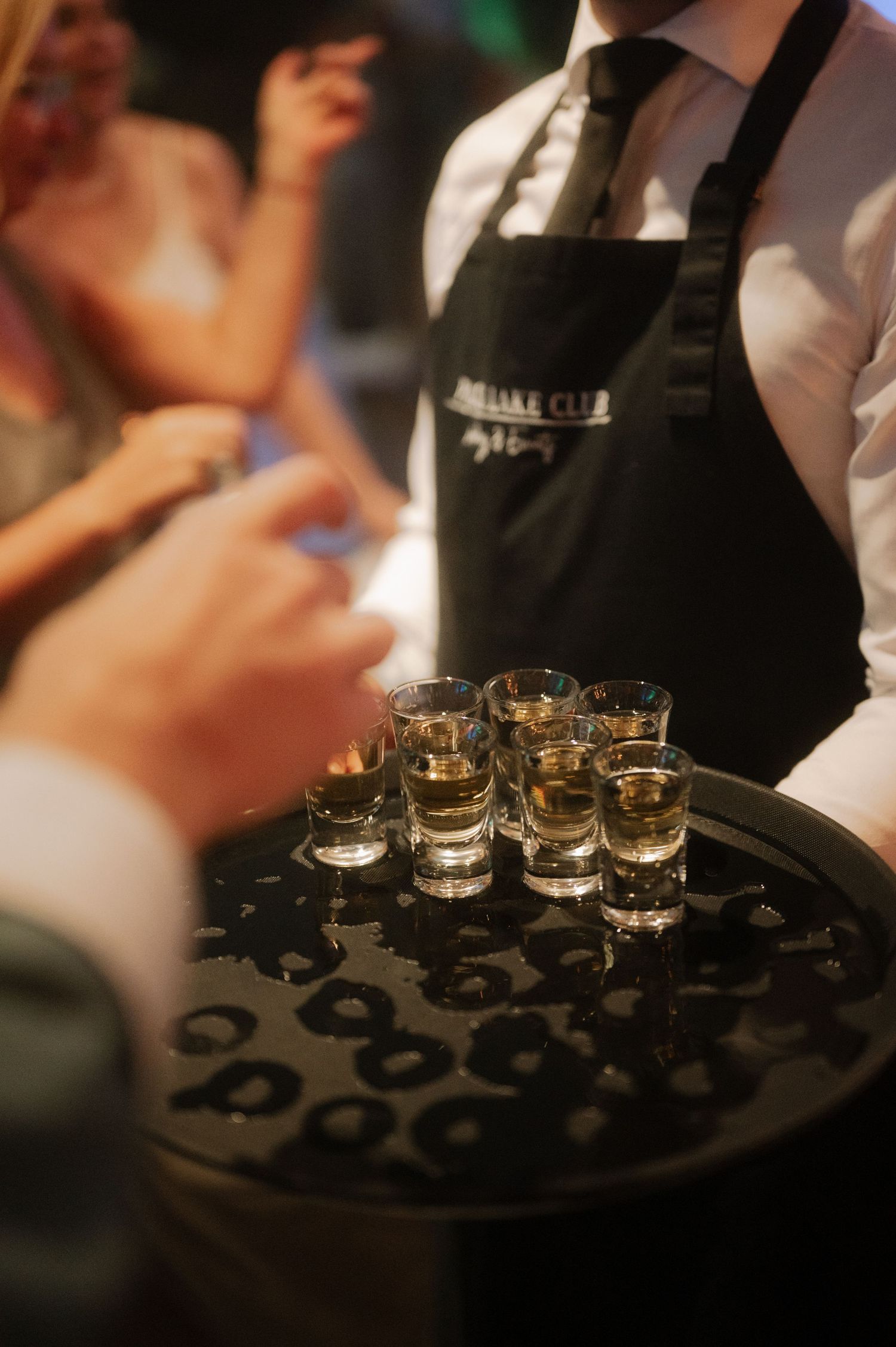 A black serving tray holds multiple shot glasses filled with clear liquid being served at an event.