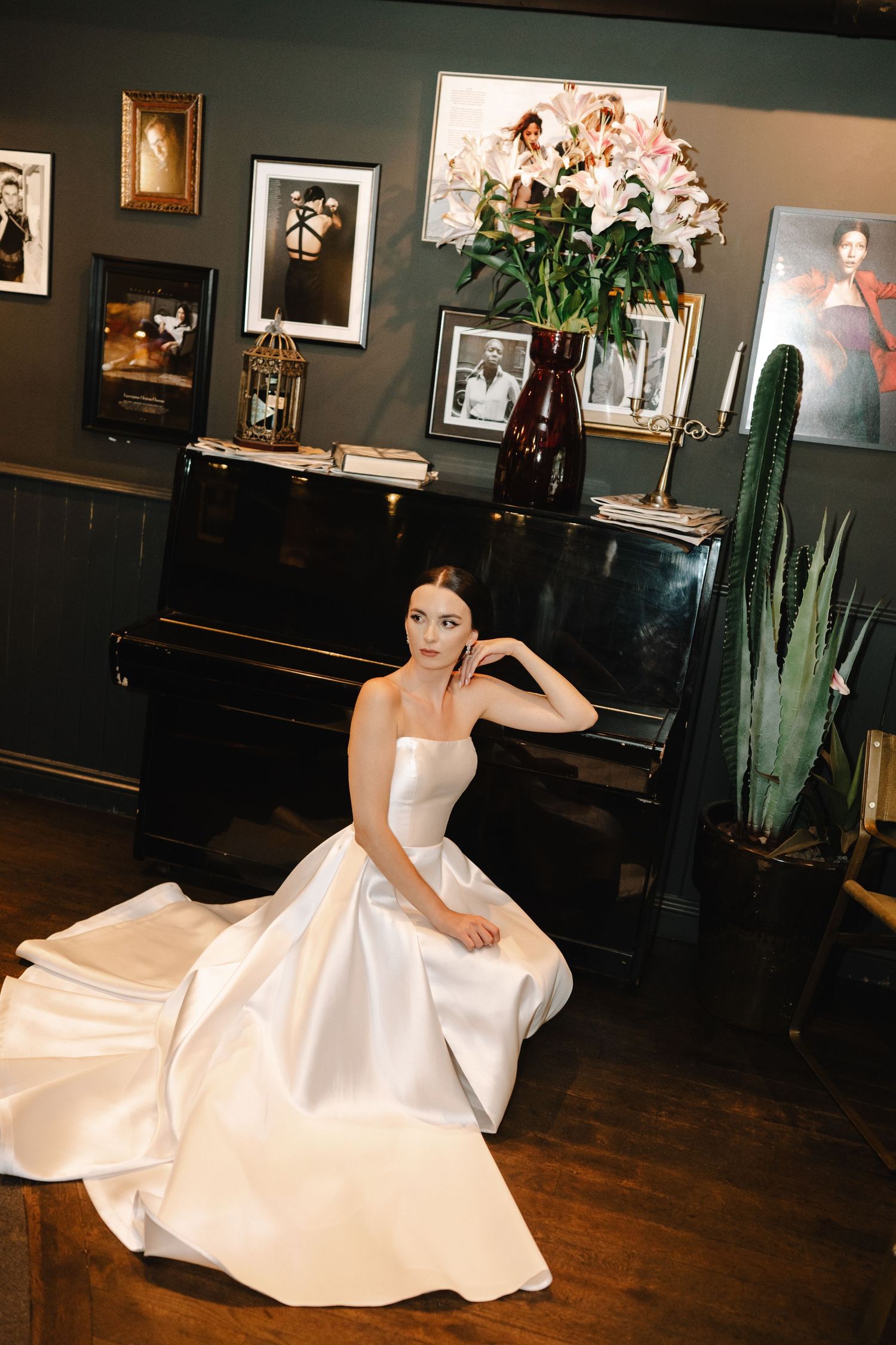 An elegant white wedding gown with a flowing skirt is displayed in a room with a black piano and potted cactus.