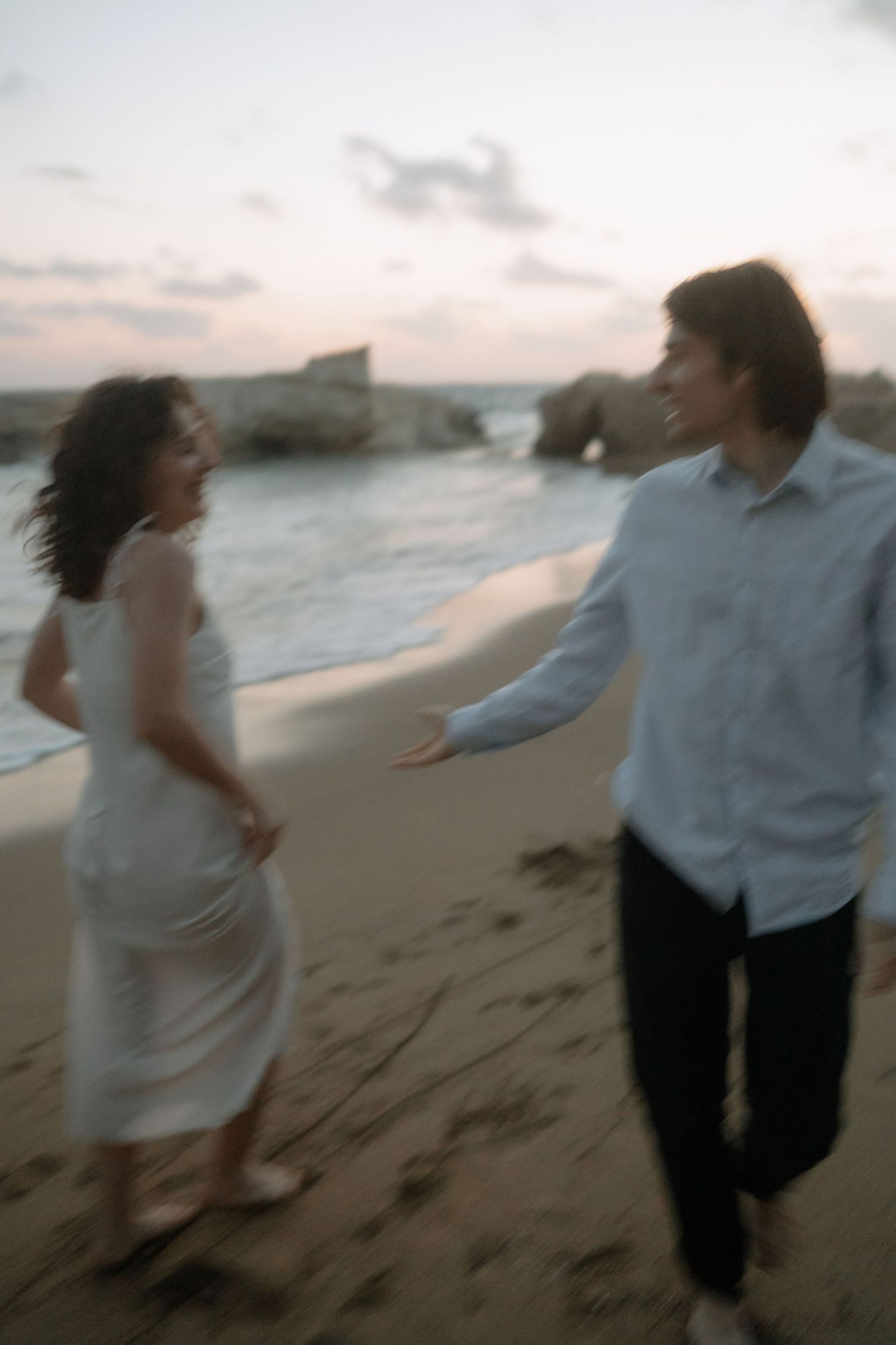 A couple walks hand in hand on a sandy beach at dusk, with ocean waves and rocks in the background.