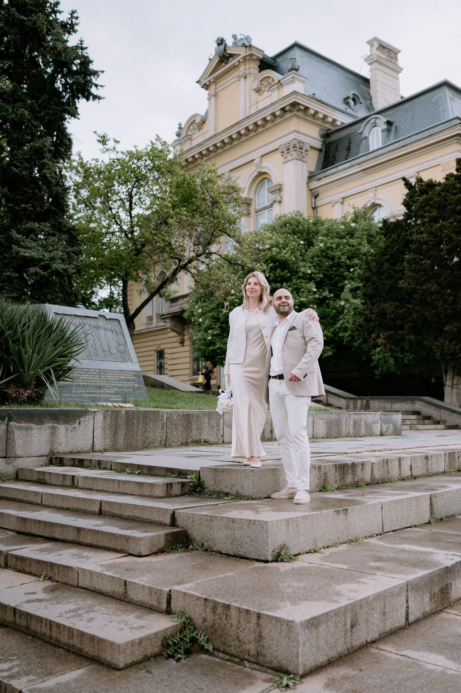 Two people in white outfits pose on stone steps with ornate building in background.