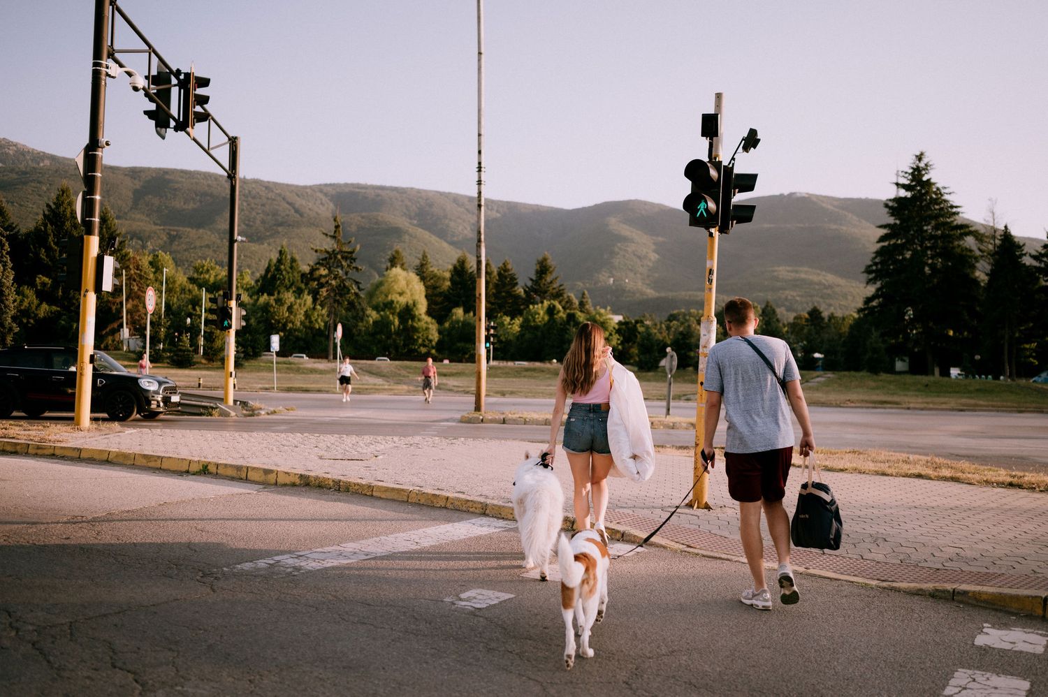 A couple walks their white dog on a paved path with mountains in the background.