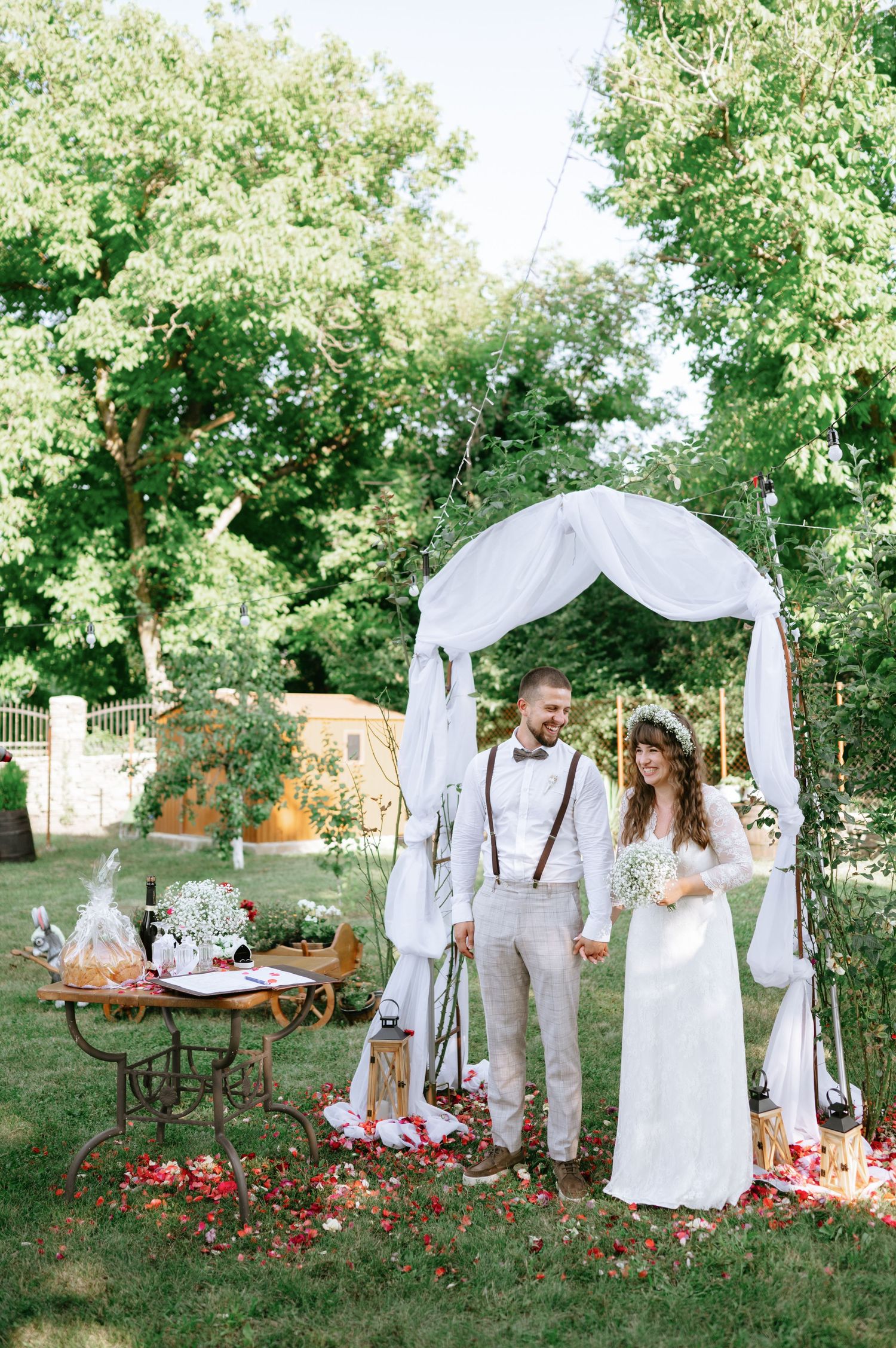Summer garden wedding setup featuring white arch, antique furniture and red flower petals on lawn.