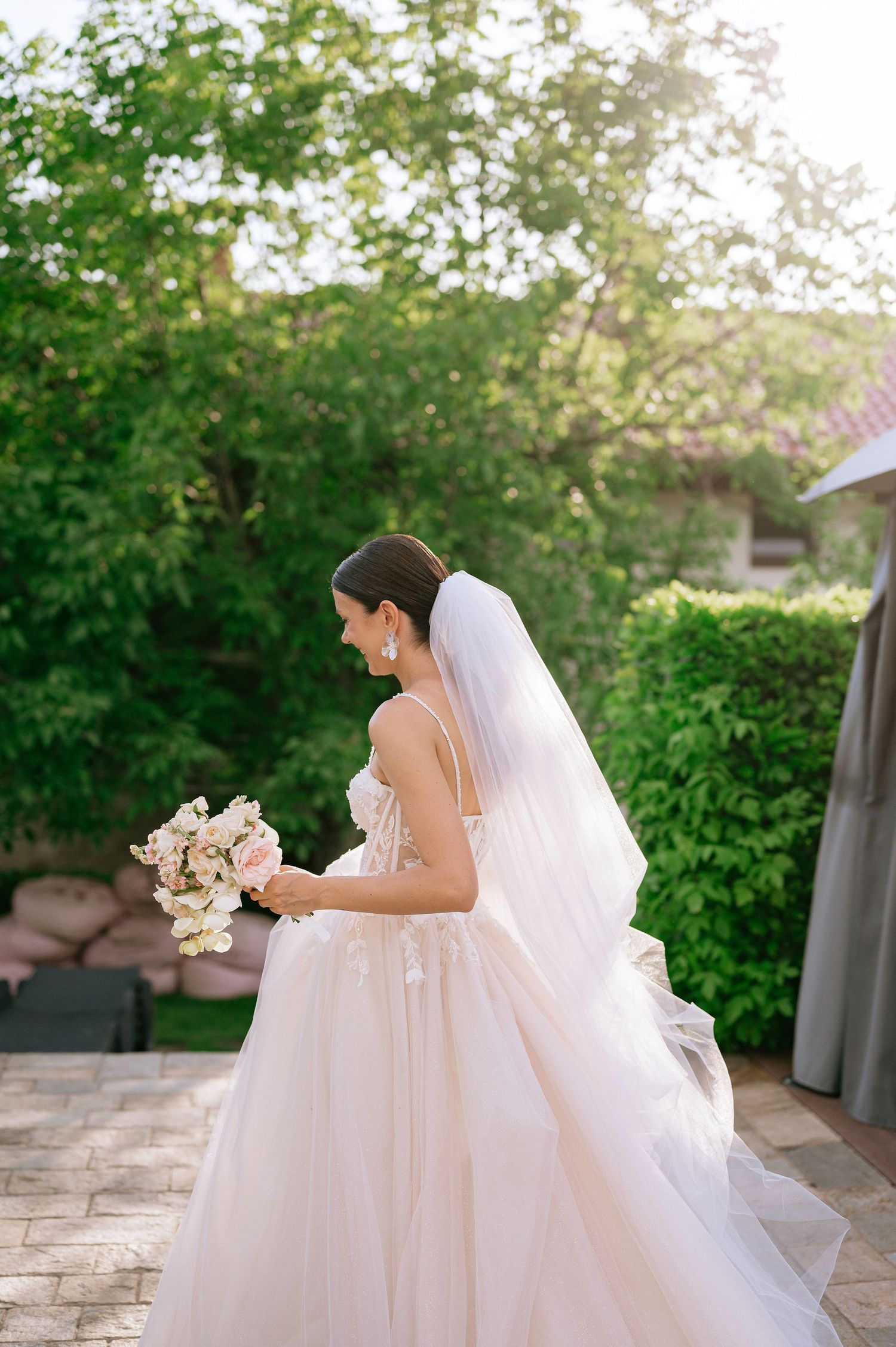 Bride in a flowing tulle gown holds a blush bouquet while walking on stone patio.